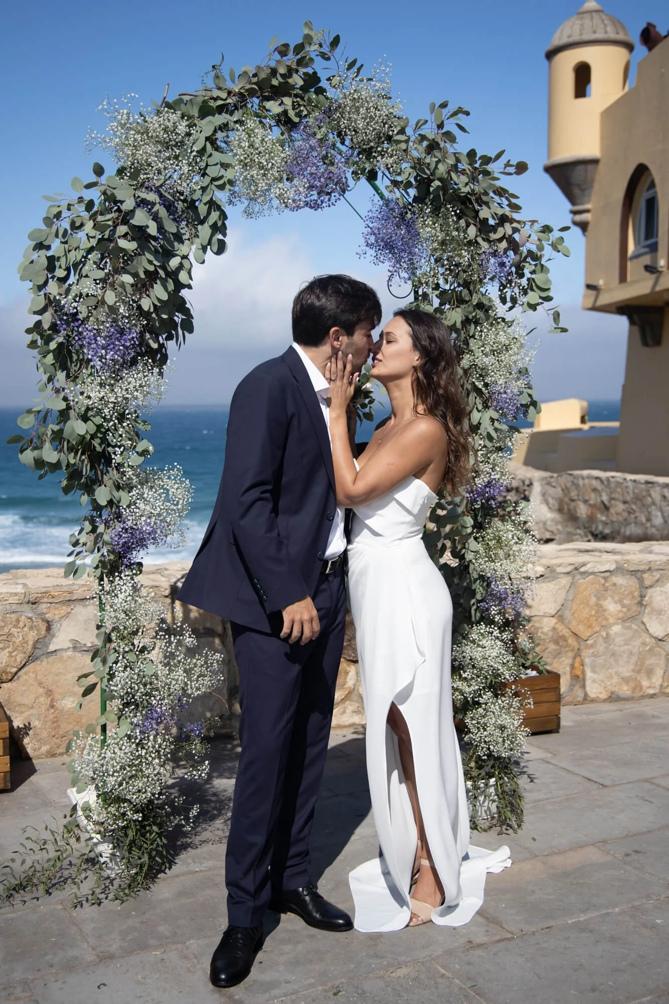 A wedding couple stands face to face with eyes closed, noses touching, in front of a floral arch by the ocean. The bride is wearing a white strapless dress with a slit, and the groom is in a dark suit. There is a stone wall and a castle-like building in the background