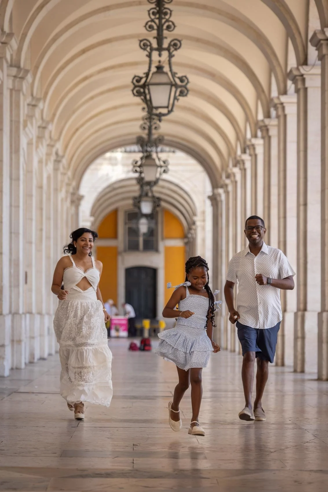 A family of three running through an arched corridor with hanging lamps. The woman is wearing a white dress, the girl in a white dress, and the man in a white shirt and dark shorts. They are smiling and appear joyful. Lisbon Family Photoshoot.
