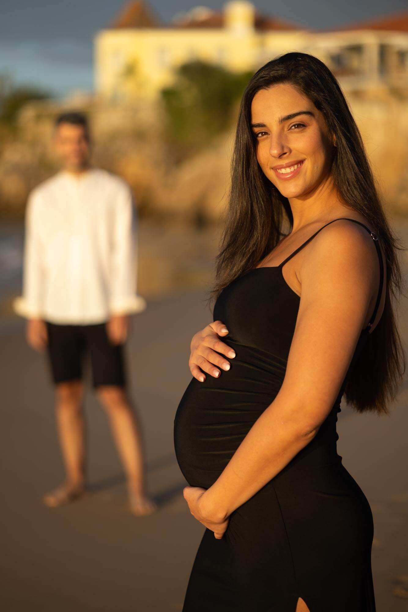 A pregnant woman in a black dress smiling at the camera with a man standing in the background on a beach during sunset.