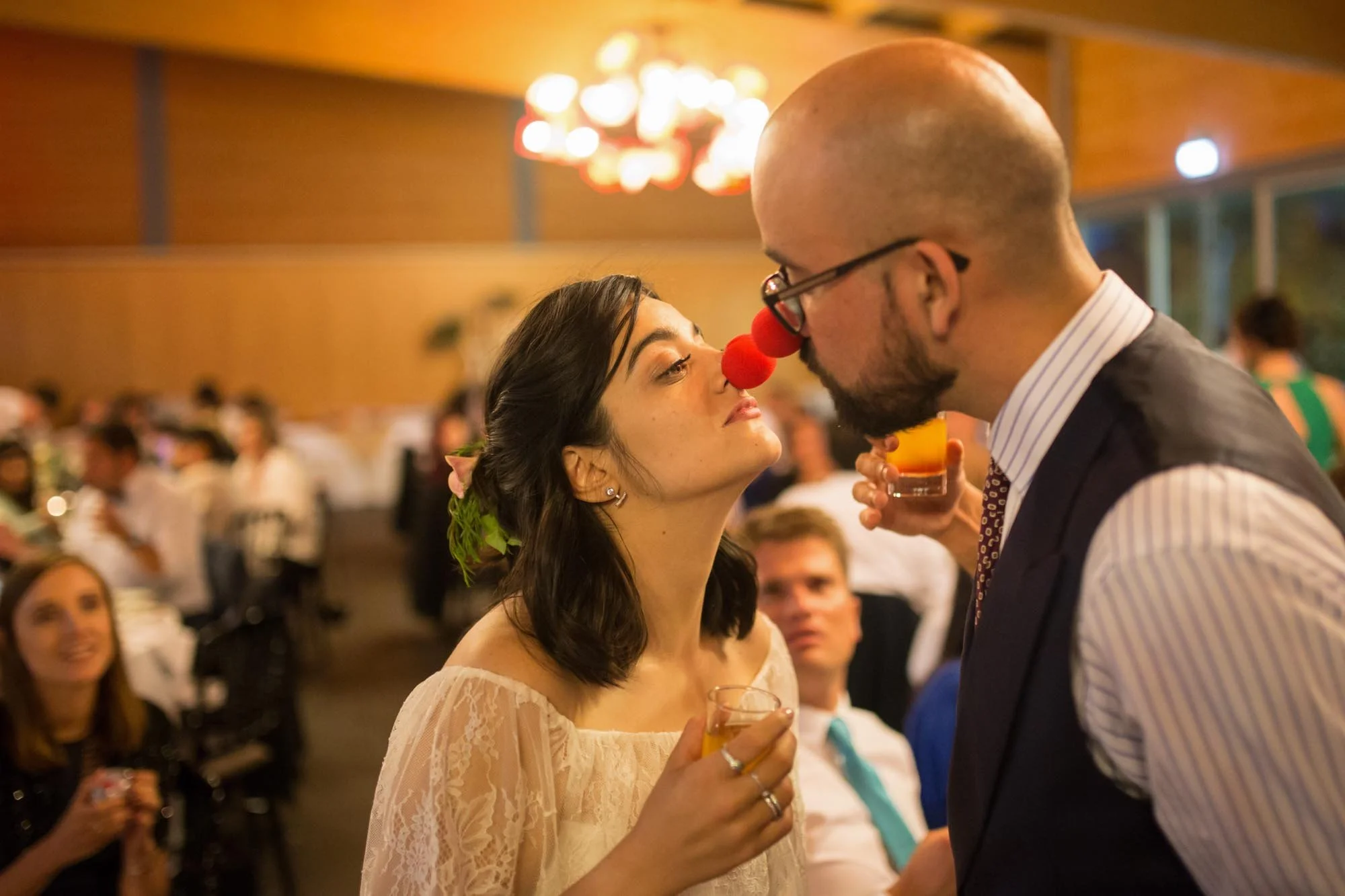 A woman and a man at a wedding or party, both with noses painted red, pretending to touch noses, with guests seated in the background.