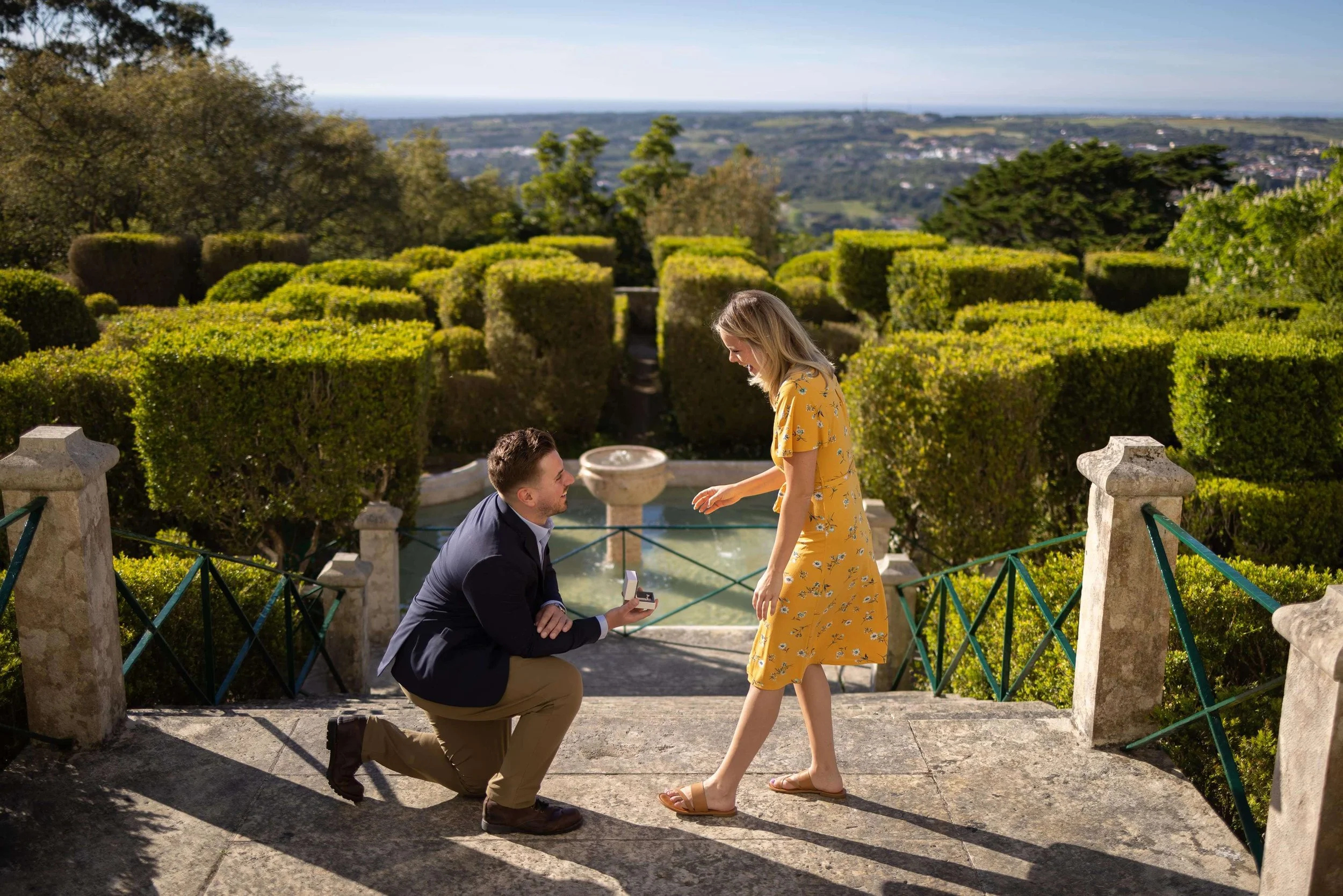 A man is kneeling on one knee proposing to a woman in a yellow dress on a stone terrace with a fountain and lush green hedges in the background.