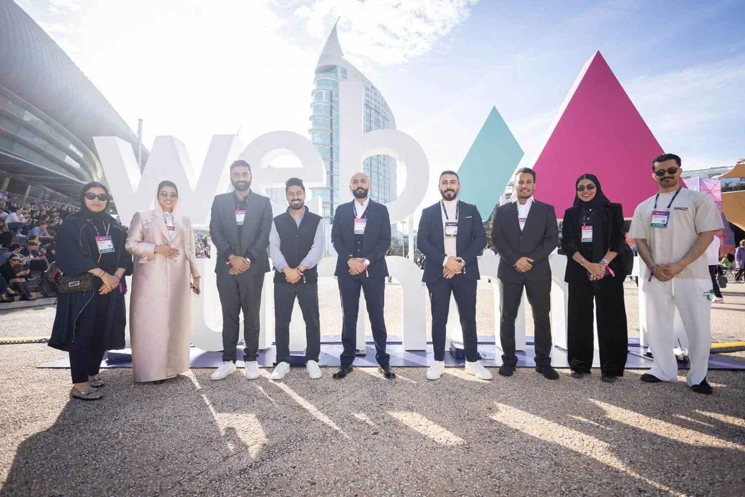 Group of ten people standing outdoors in front of large white letters spelling "web" and a colorful geometric backdrop, with modern buildings and a crowd sitting on steps behind them. They are dressed in formal and business casual attire, some wearin