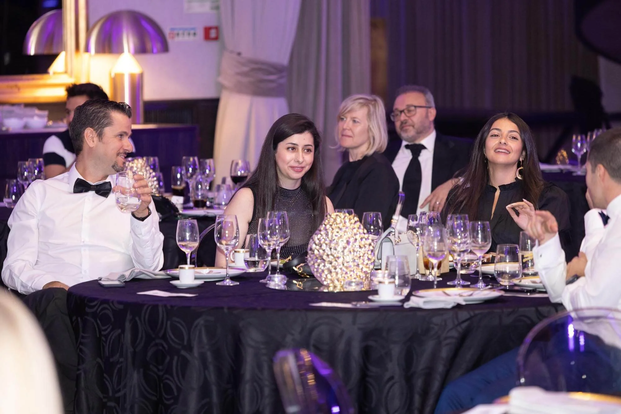 People enjoying a formal dinner at a restaurant or banquet hall, dressed in formal attire with wine glasses and a decorative centerpiece on the table.