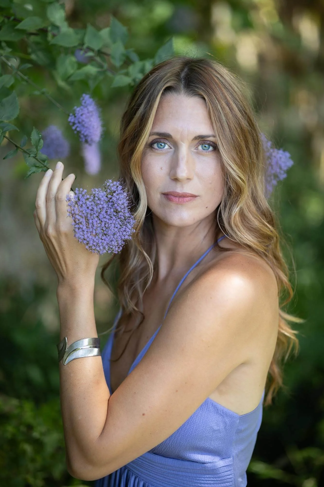 A woman with long wavy blonde hair and blue eyes wearing a light purple dress, standing outdoors among greenery and purple flowers, holding a cluster of purple flowers near her face.