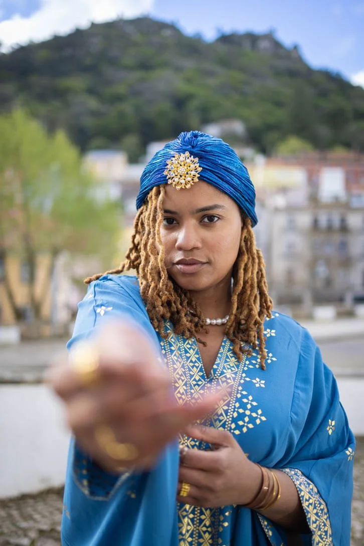 Woman with blonde dreadlocks wearing a blue headscarf and traditional clothing, pointing toward the camera outdoors with trees and a mountain in the background.