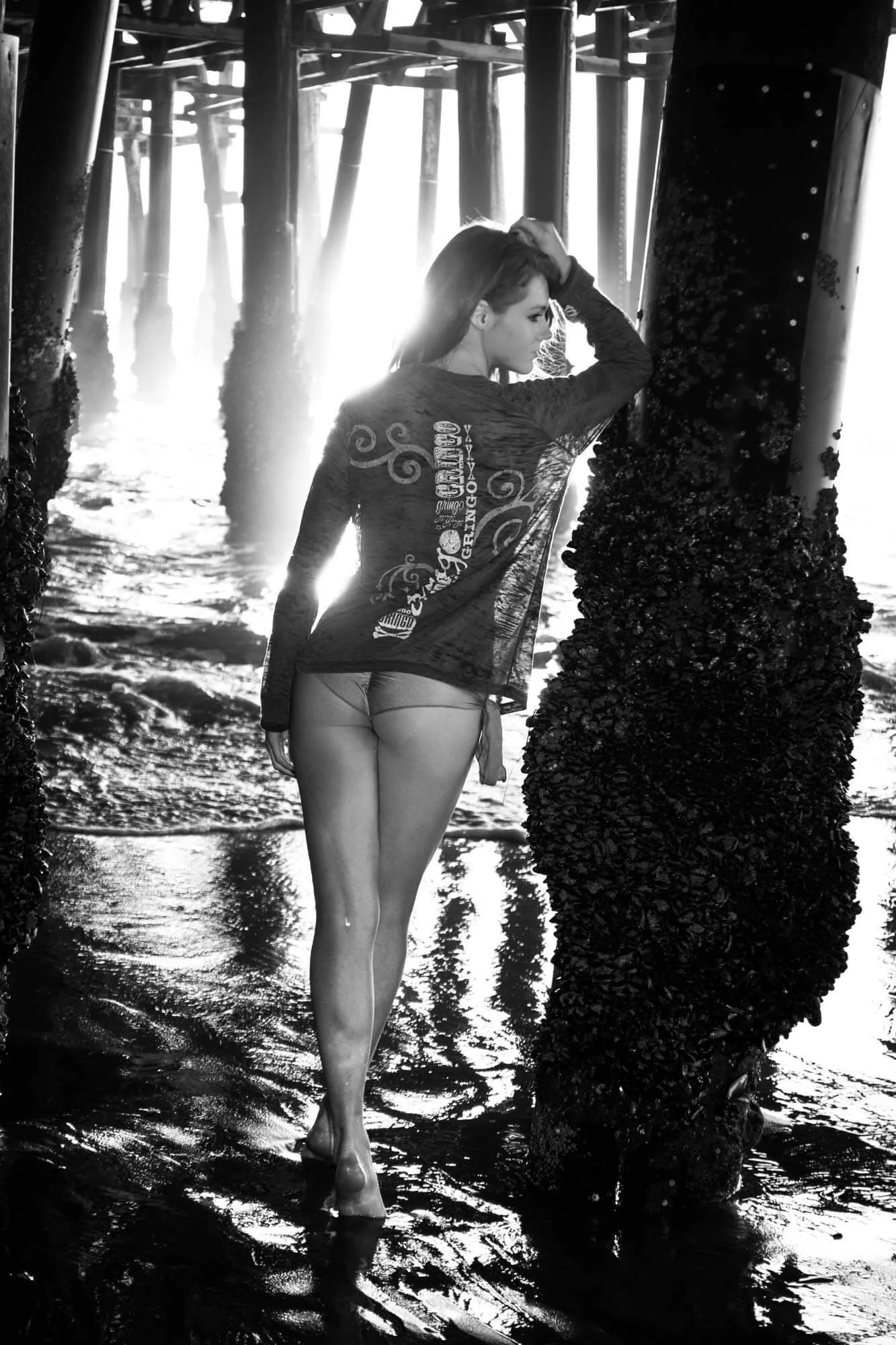 A woman standing barefoot in water under a pier, leaning against a barnacle-covered post, with sunlight shining behind her.