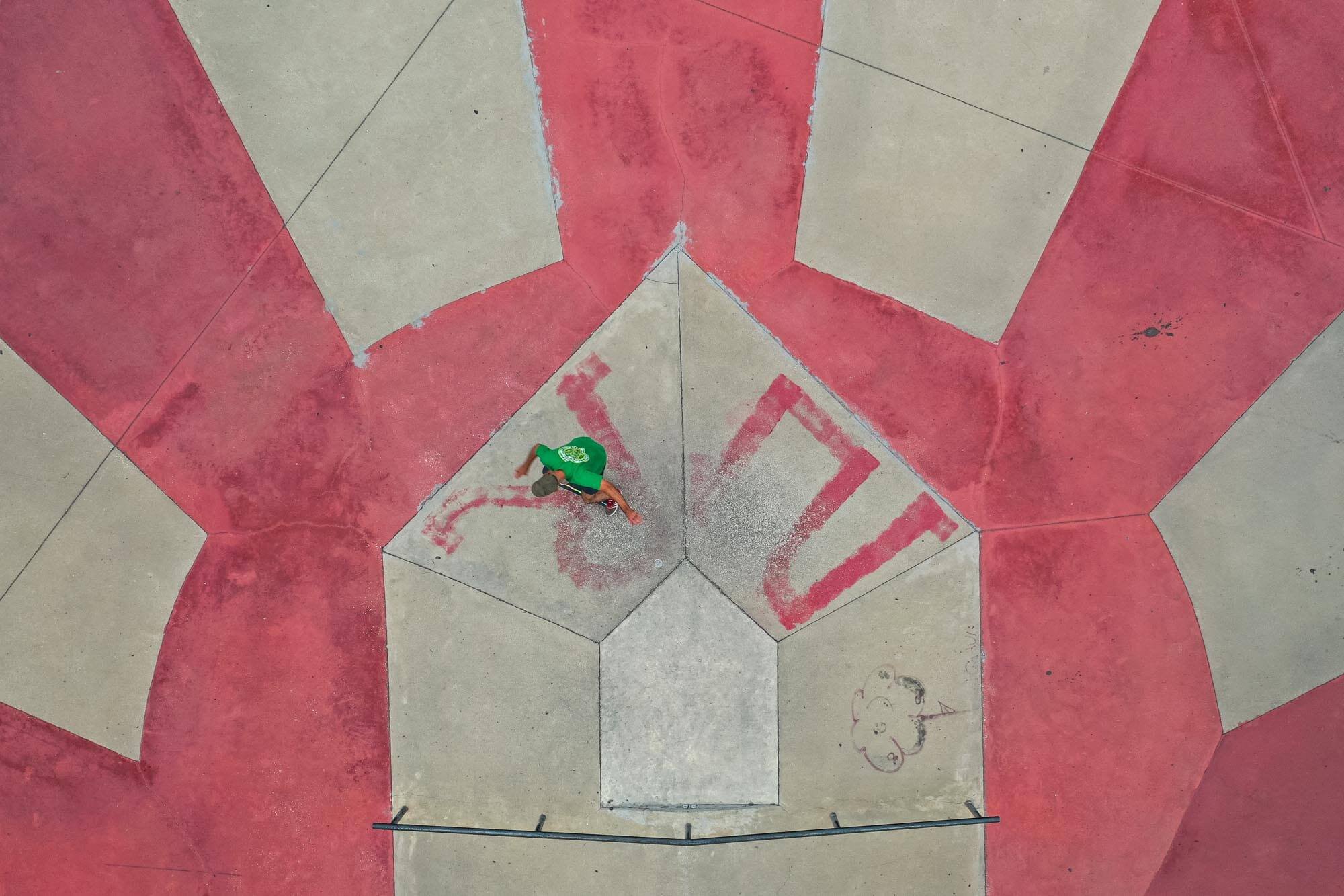 A person wearing a green shirt and black shorts is walking on a concrete surface with red and beige painted geometric shapes. The photo is taken from above, showing the person near a red triangular shape with some graffiti.