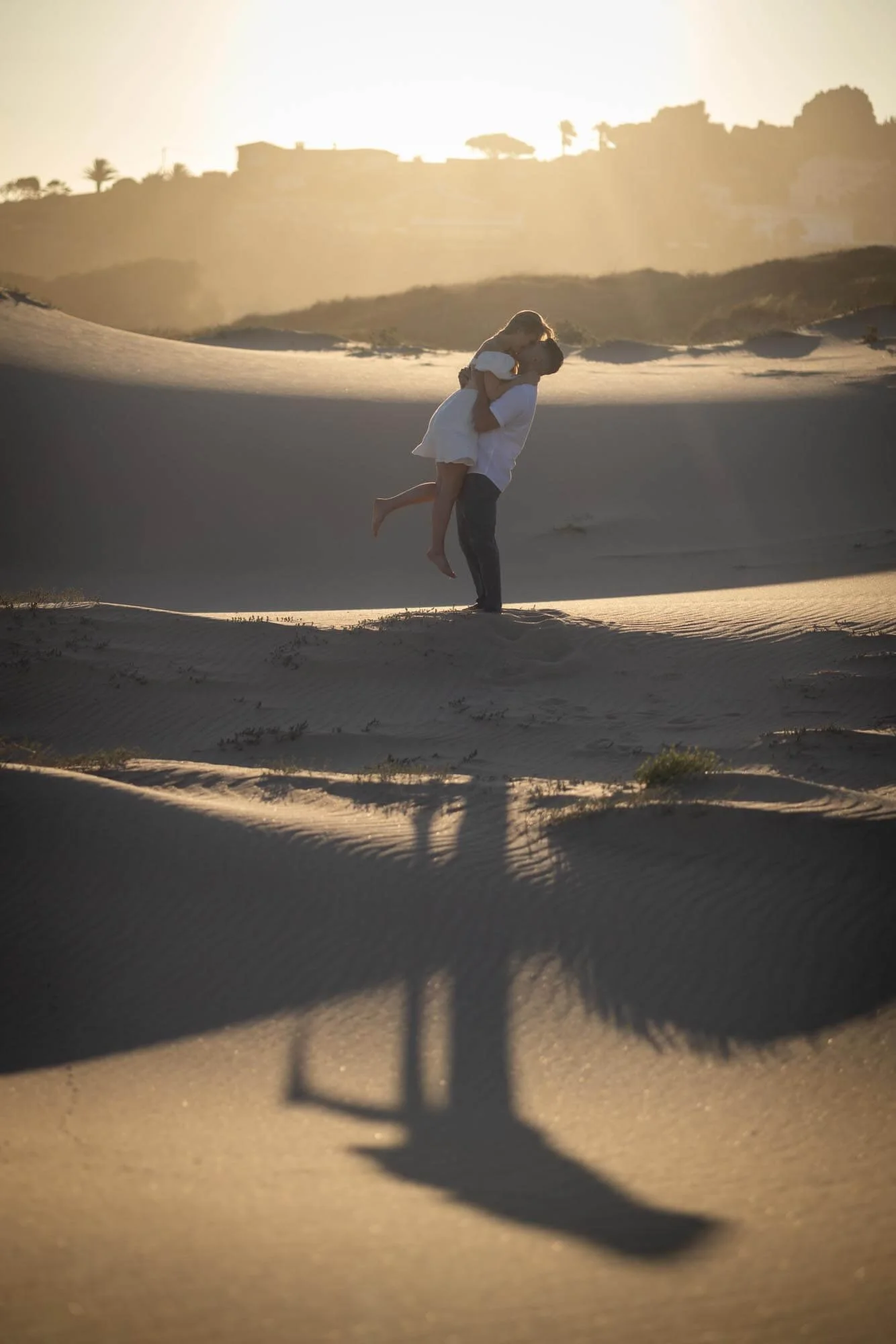A couple in a desert with sand dunes, with the man holding the woman in his arms as the sun sets behind them.
