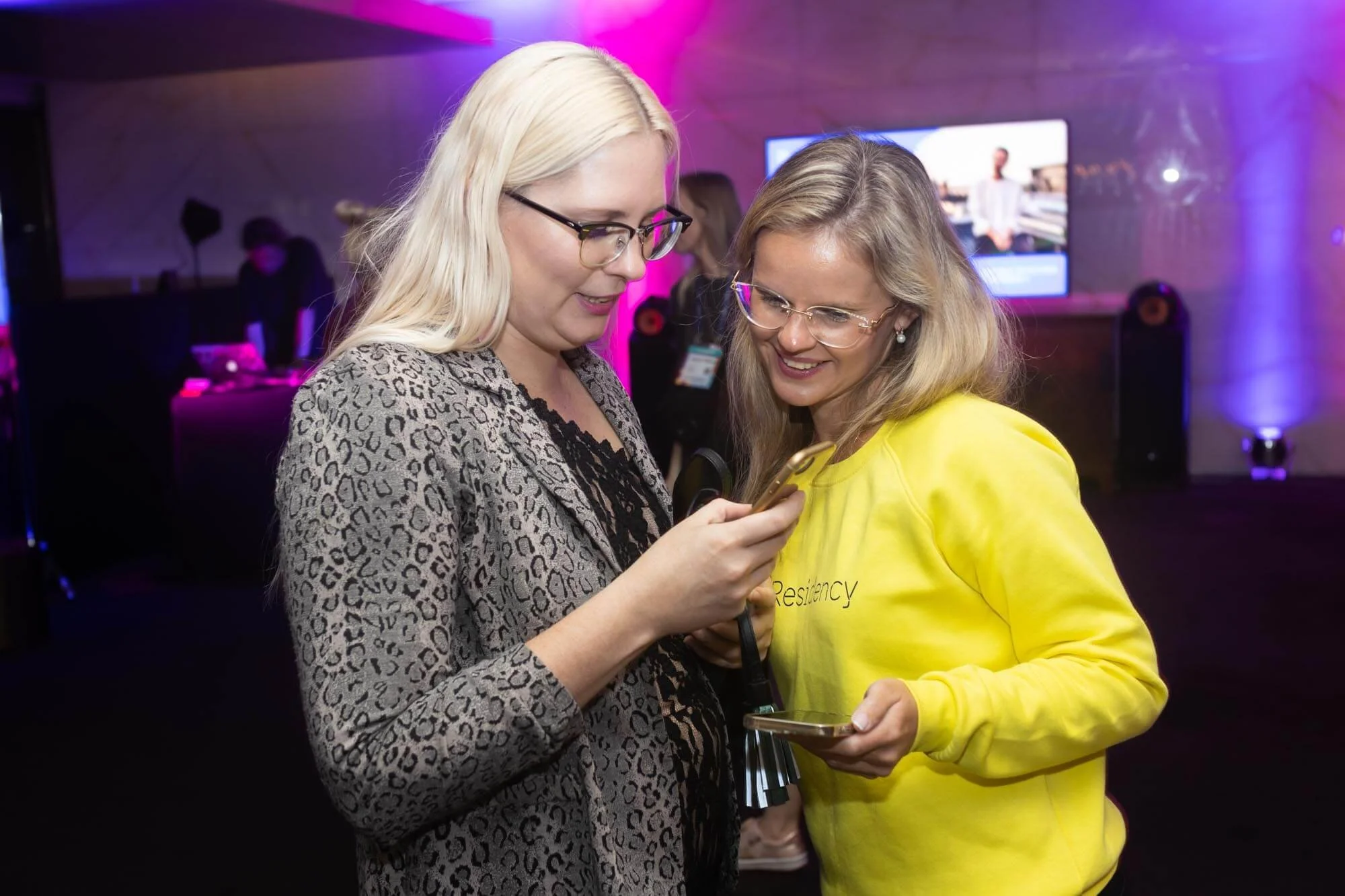Two women with glasses looking at a smartphone and smiling at a social event, with colorful lighting and a large screen in the background.