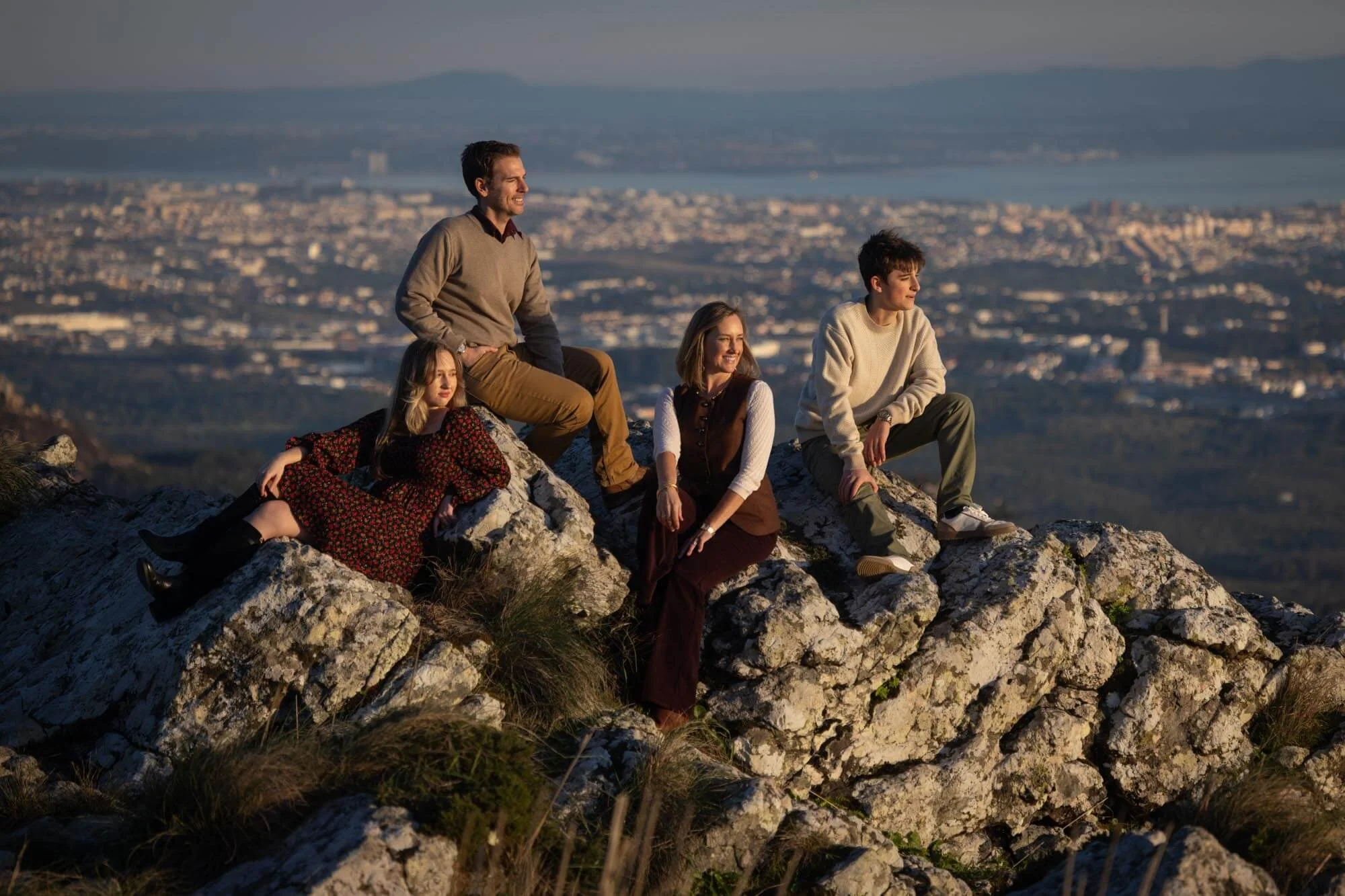 Five people sitting on rocks on a mountain summit during sunset, with a cityscape and water body in the background.
