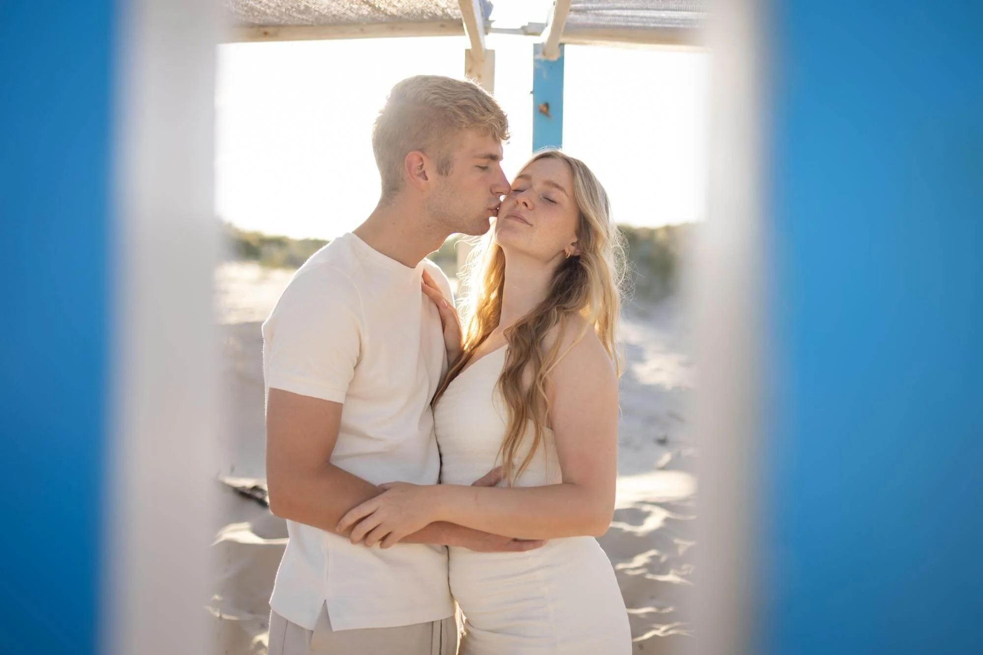 A young couple stands on a beach facing each other, with the man kissing the woman on the forehead as she closes her eyes. They are framed by the opening of a lifeguard stand painted in blue and white, with sunlight illuminating the scene.