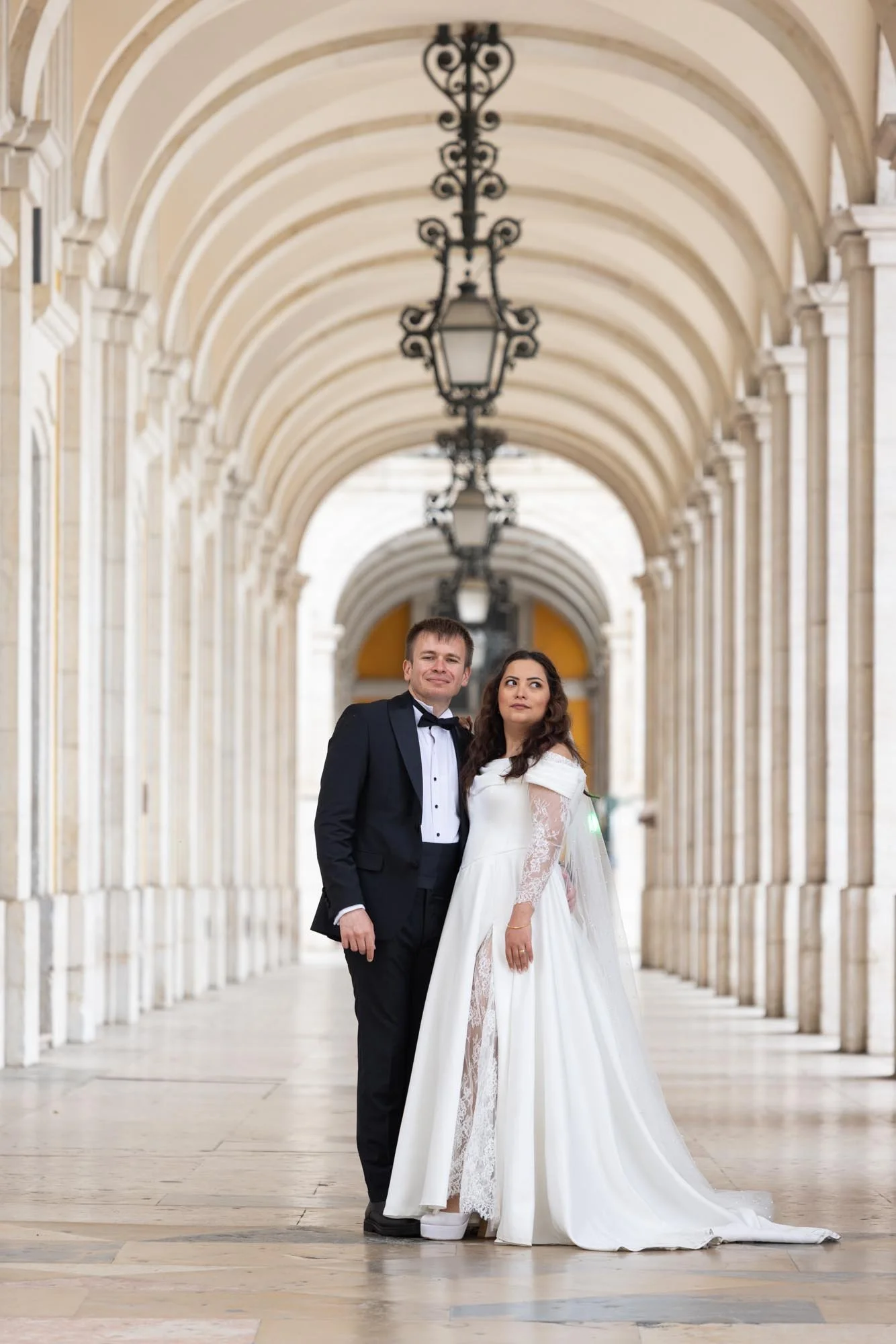 A bride and groom in wedding attire standing in an elegant arched hallway with hanging lanterns.