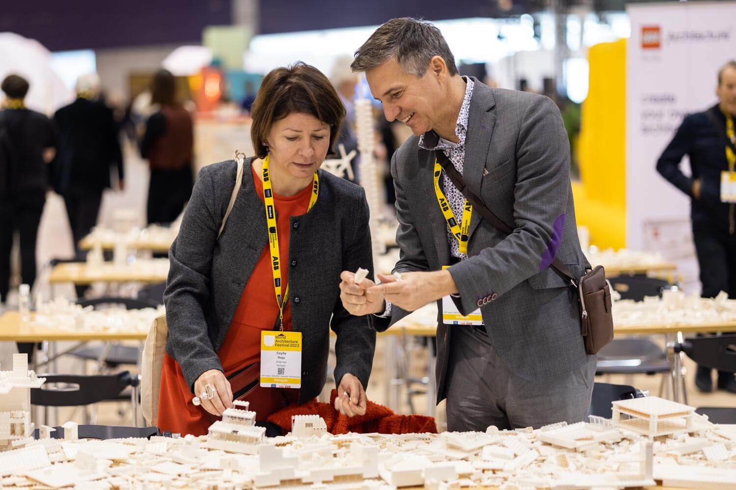 Two people, a woman and a man, are viewing a scale model of white buildings at an indoor exhibition. Both wear yellow lanyards, and the background features other attendees and display booths.