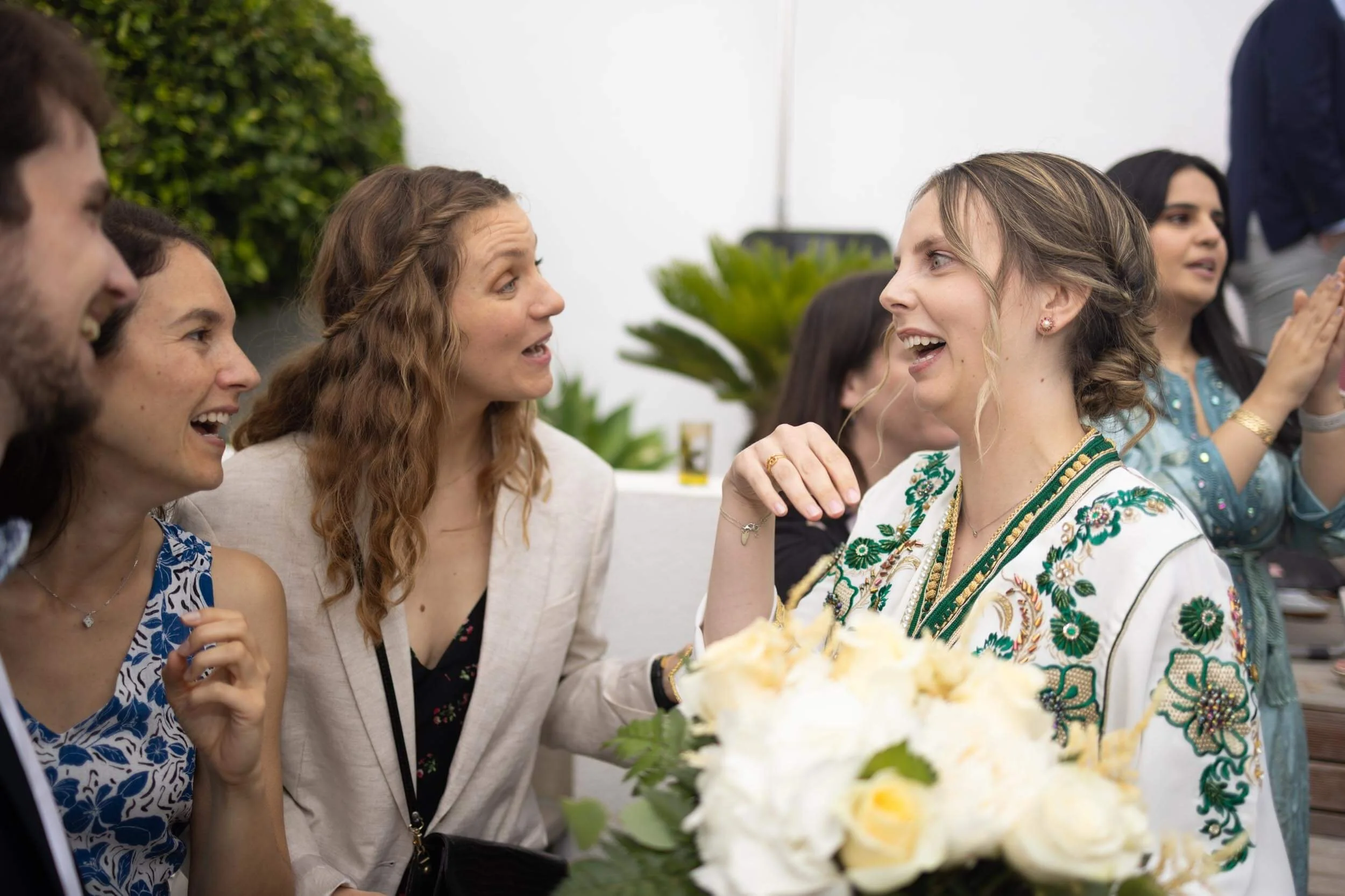 Group of people celebrating at a social event, smiling and engaging in conversation with a woman wearing a white embroidered dress, flowers in the foreground, and a lush green plant in the background.