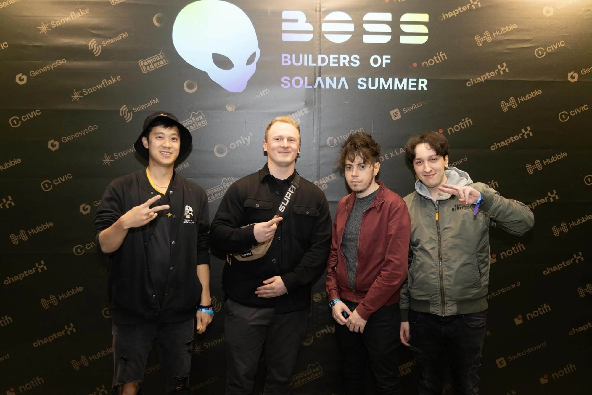 Four young men standing in front of a black backdrop with logos and text, including a large white alien head, 'BOSS' in large letters, and the phrase 'BUILDERS OF SOLANA SUMMER'. All four are smiling, with the first on the left flashing a peace sign,