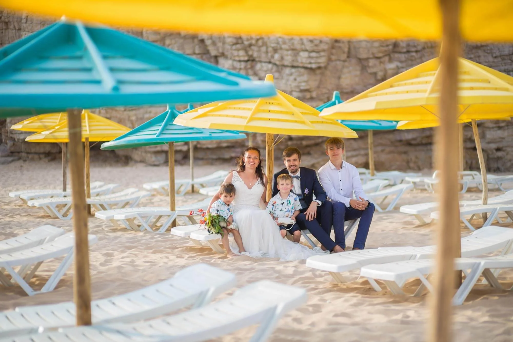 A family sitting on a chair under yellow and blue umbrellas on a sandy beach with stone cliffs in the background, dressed in wedding attire.