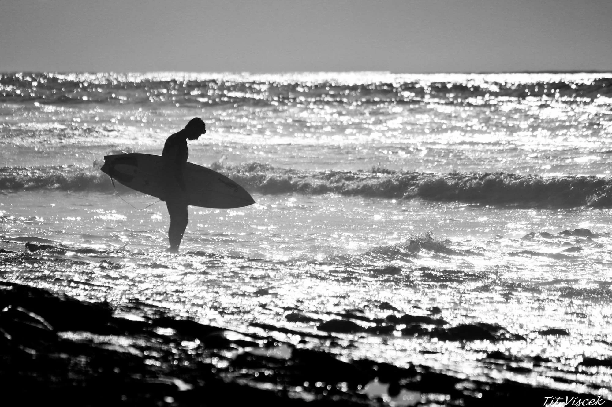 Silhouette of a surfer walking in the ocean at sunset, carrying her surfboard under her arm, with shimmering water and waves in the background.