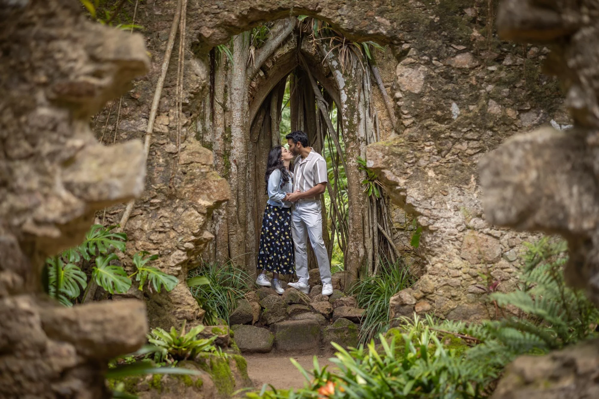 A couple stands close together, sharing a kiss under a stone archway with climbing roots, surrounded by lush green plants.