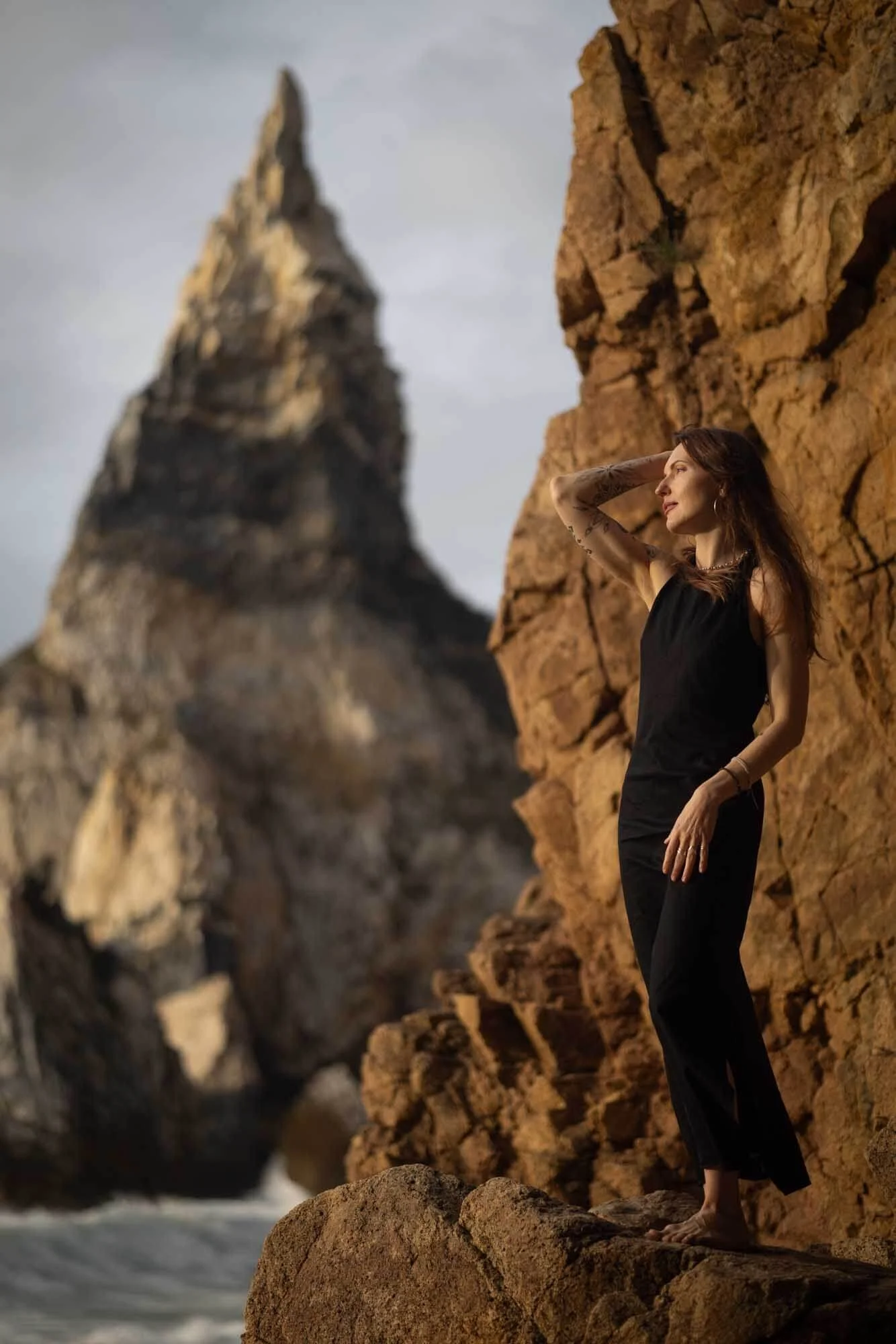 A woman standing on a rocky cliff by the ocean, with towering rock formations in the background during sunset or sunrise.