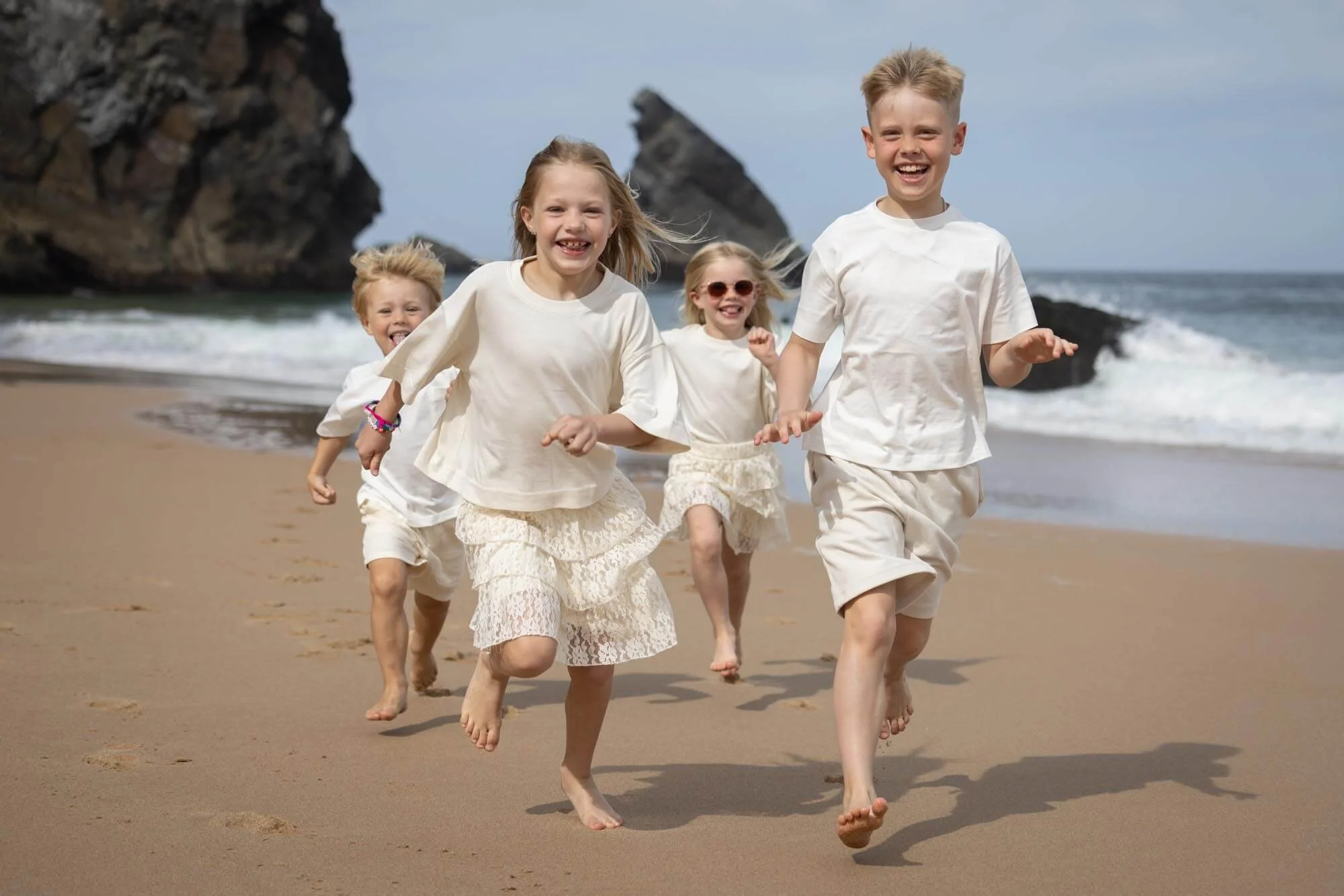 Group of five children running and laughing on the beach near the ocean with rocks in the background.