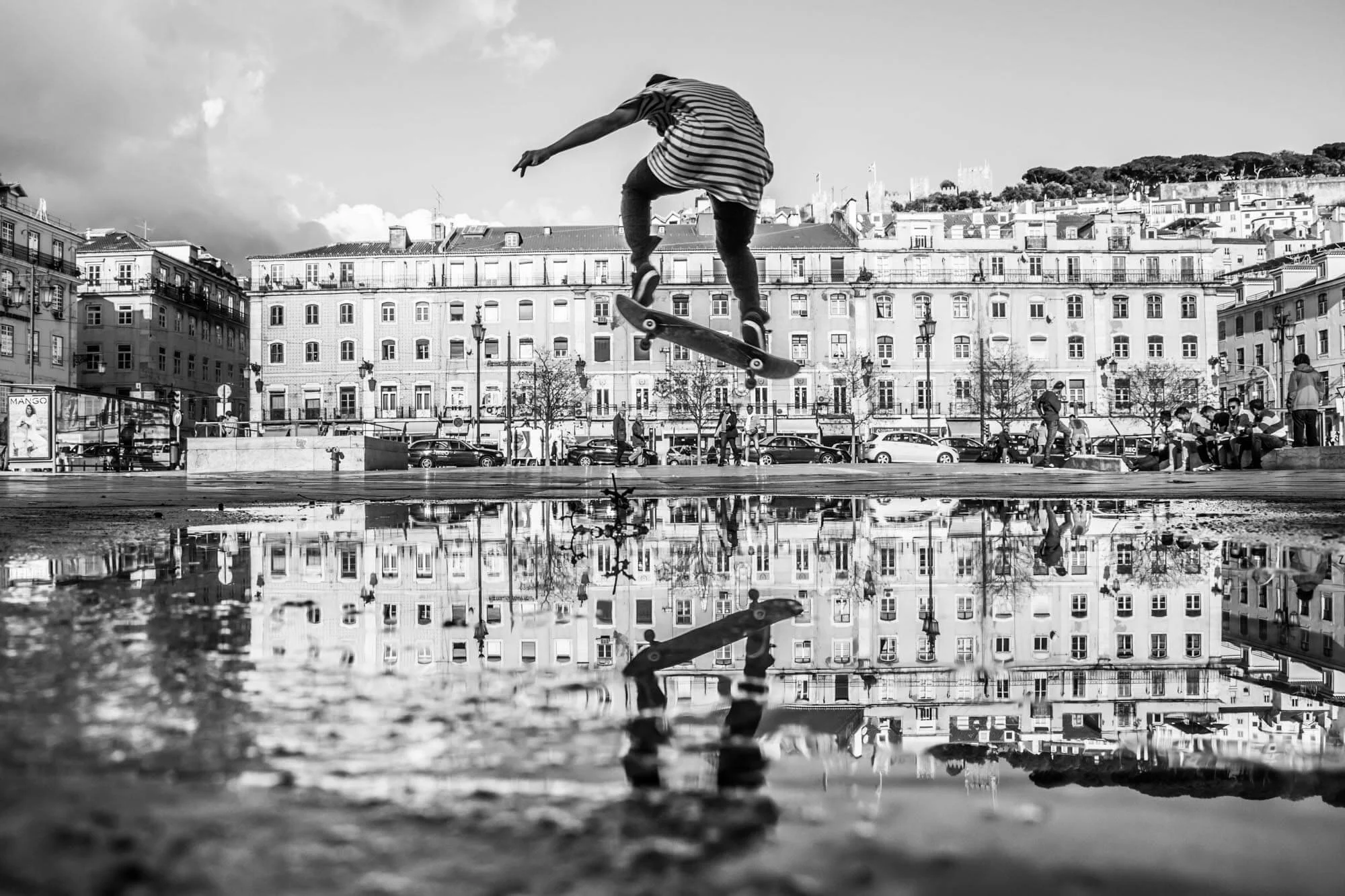 A skateboarder performing a jump in a city square with buildings in the background, reflected in a puddle on the ground.