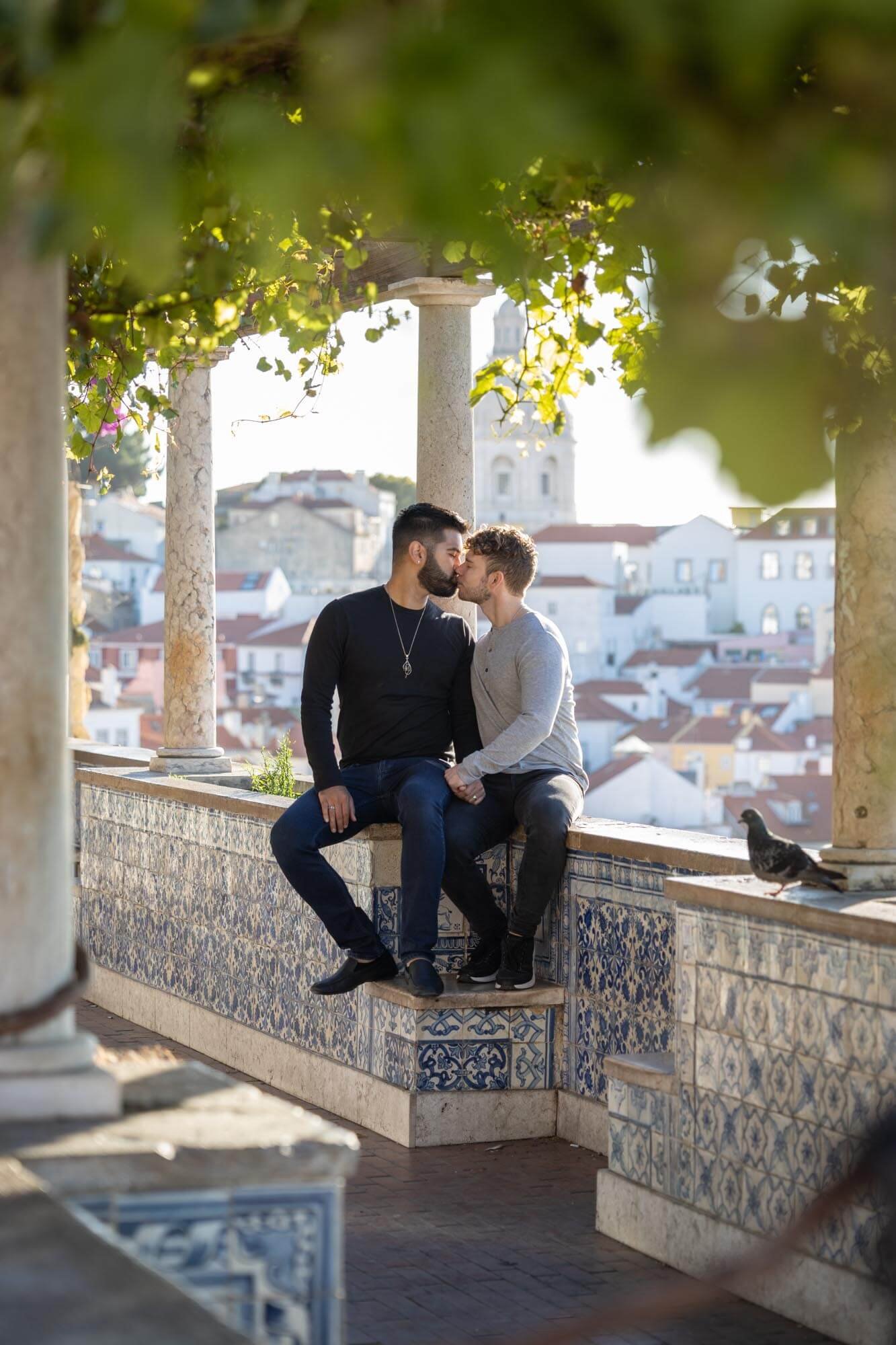 Two men sitting on a tiled ledge kissing, with a view of rooftops and a bell tower in the background, framed by leafy greenery.