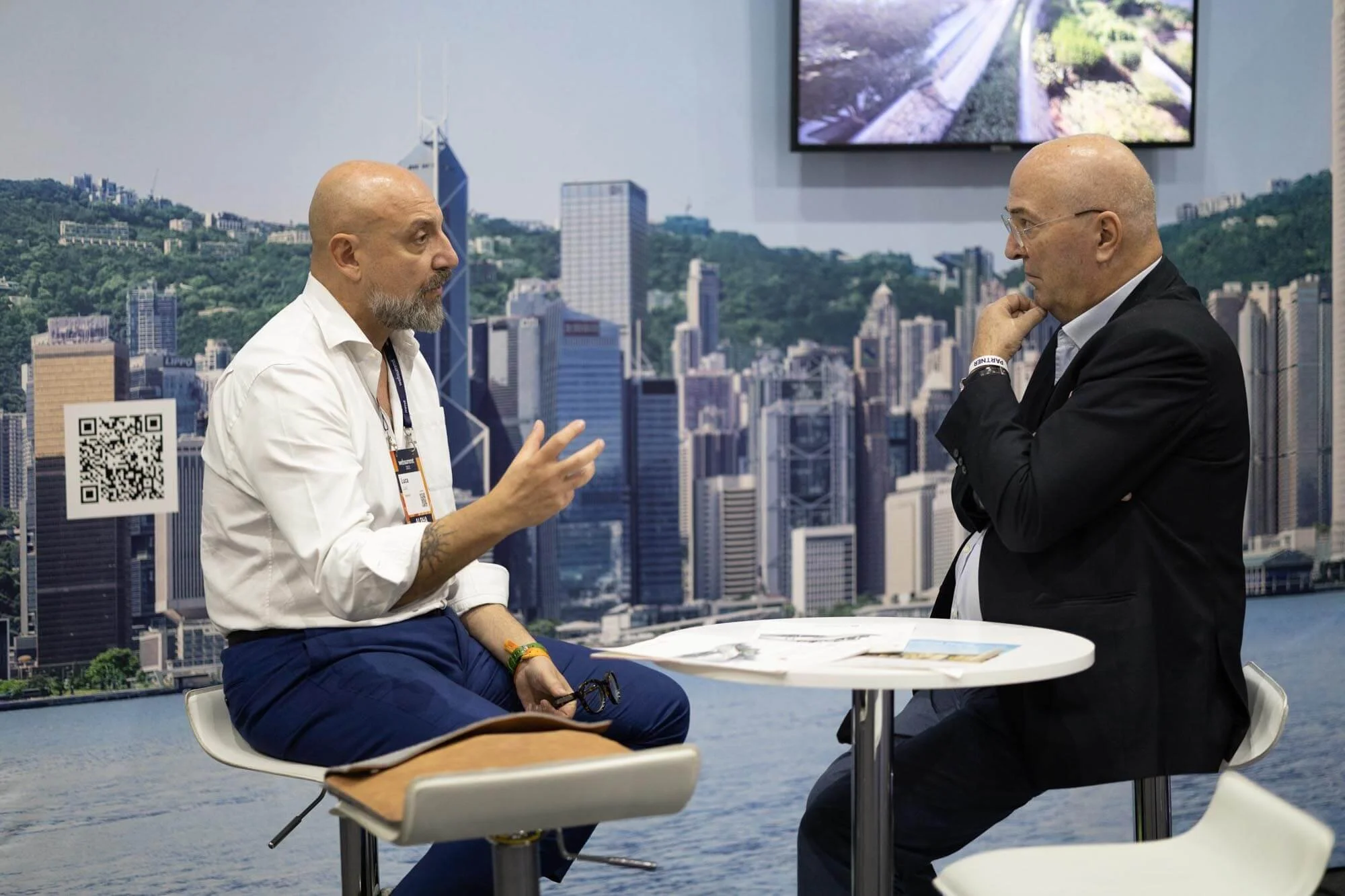 Two men engaged in a conversation, seated at a small round table with papers on it, against a cityscape backdrop with tall buildings, a river, and a large screen overhead displaying an aerial view of a park or garden.