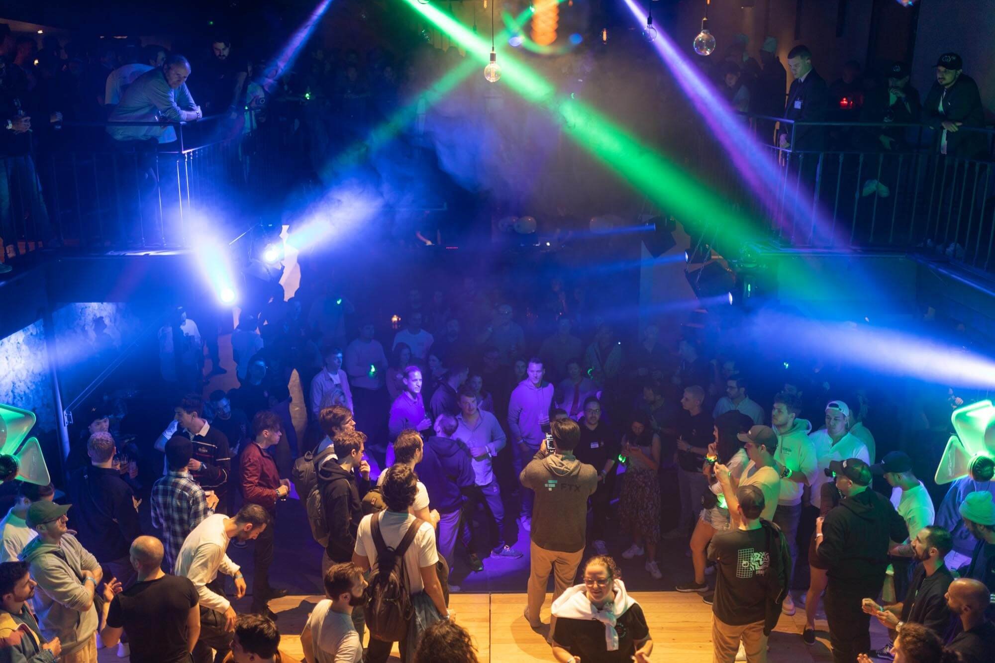 Crowd of people at a nightclub dancing under colorful laser lights and fog, with some standing on an upper balcony
