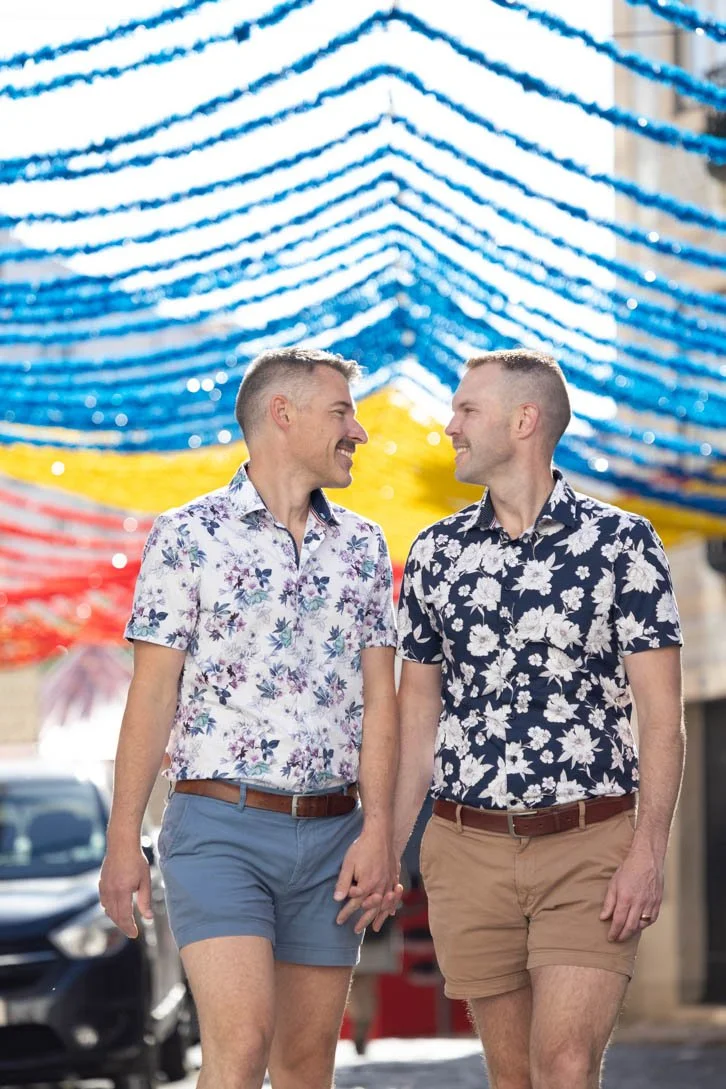Two men wearing floral shirts and shorts, holding hands and walking outdoors under colorful streamers of Santo Antonio festivel in Alfama area.
