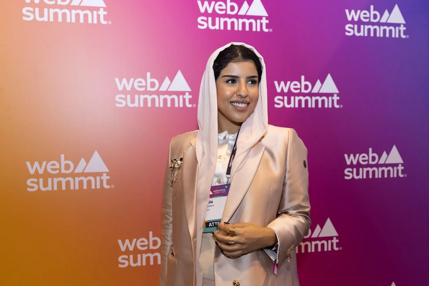 A woman in a beige blazer with a headscarf, smiling, at a Web Summit event with a colorful background featuring the Web Summit logo.