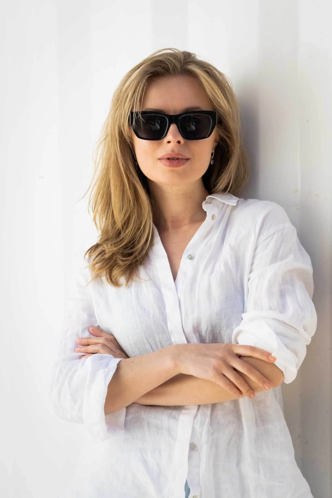 Portrait session with a beautiful blonde woman with sunglasses and white linen shirt on a white wall.