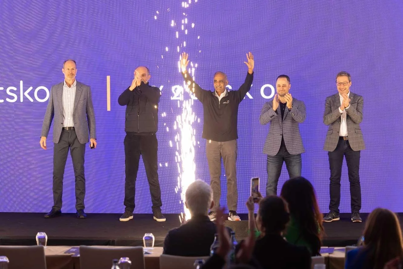 Five men standing on a stage during an award ceremony or event, with a large blue backdrop displaying 
