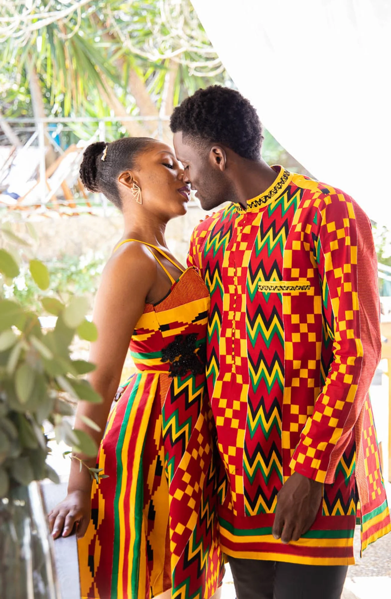 A couple dressed in traditional African kente cloth, standing close with foreheads touching, outdoors with greenery in the background.
