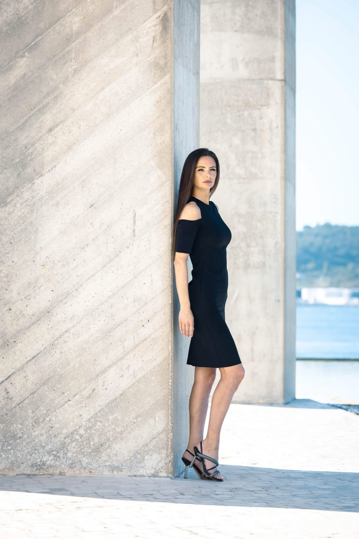 A woman in a black dress and high heels stands outdoors leaning against a concrete wall near a body of water, with a mountain in the background.