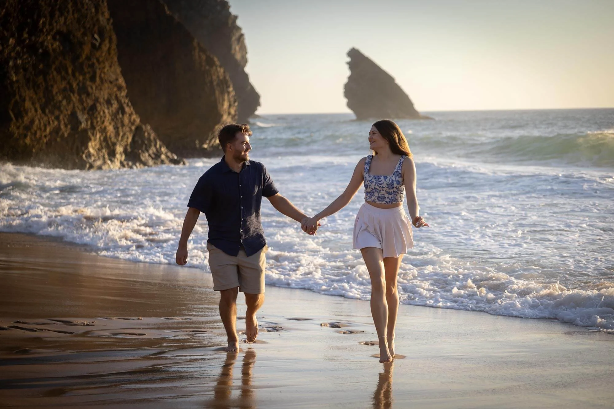 A couple walking hand in hand along the beach during sunset, with rocky cliffs and the ocean in the background.