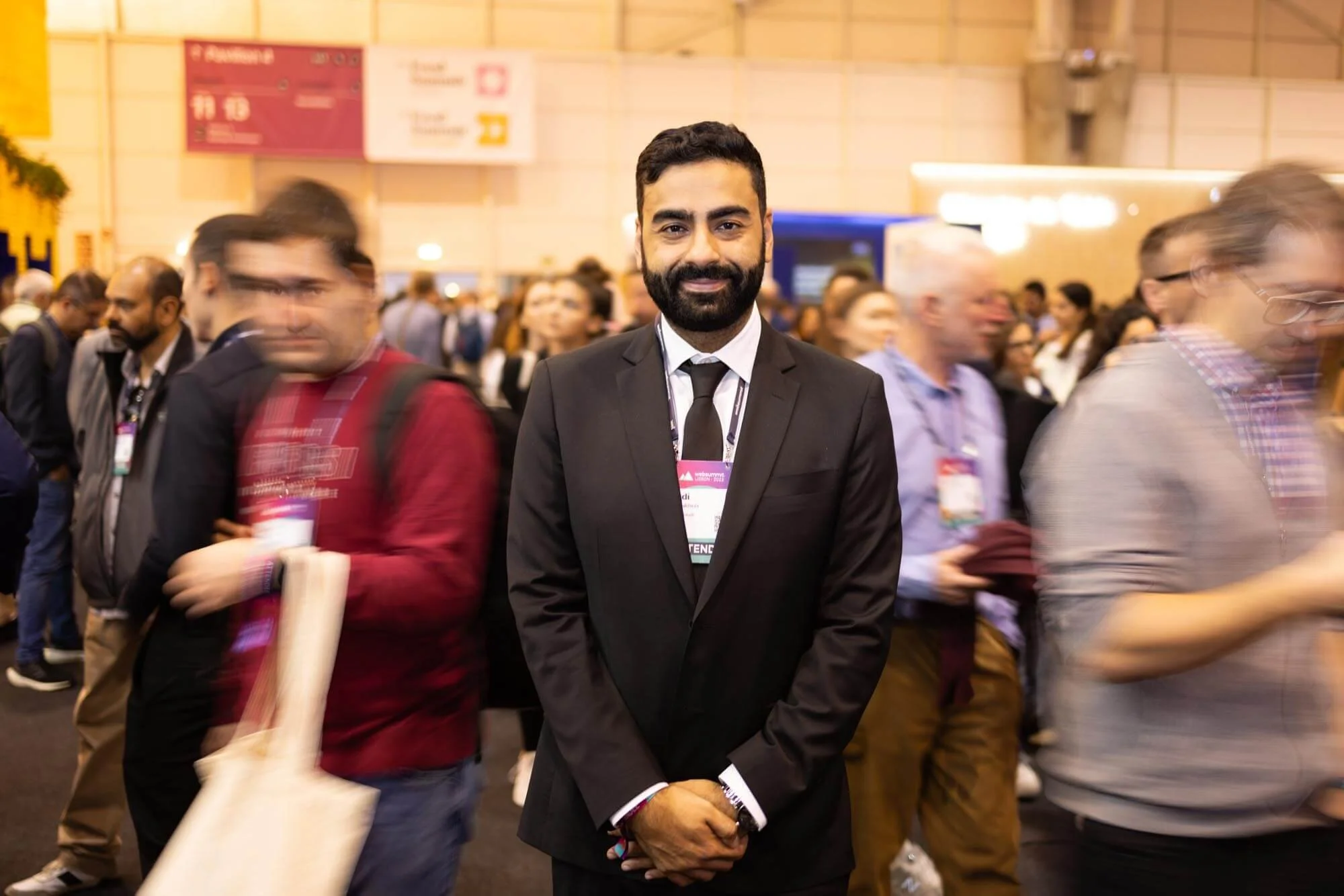 A man in a black suit and tie standing with his hands clasped in front of him, smiling at the camera, surrounded by a bustling crowd at an indoor event.