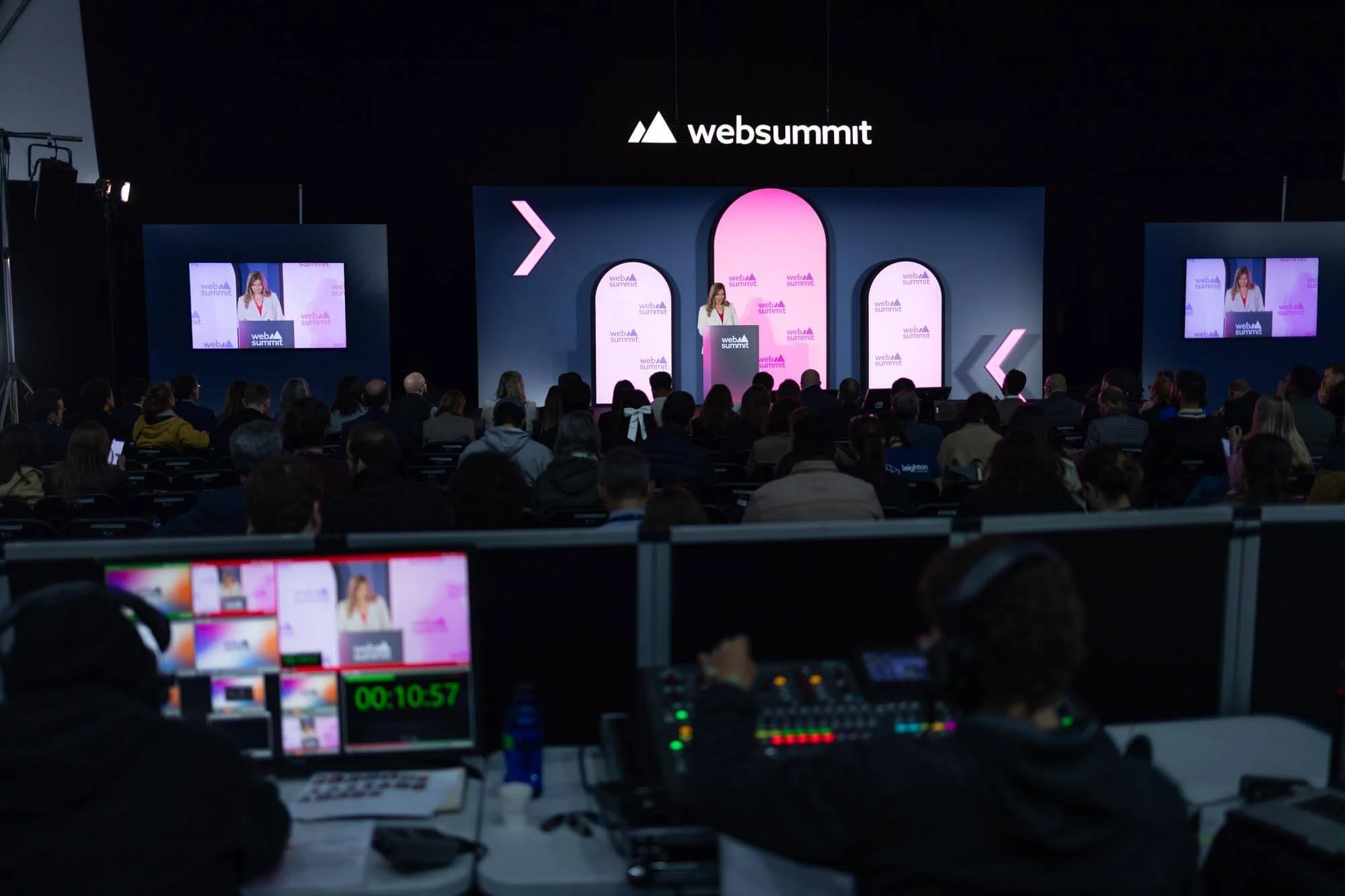 A woman speaking at a podium during a conference at WebSummit, with an audience in attendance and multiple screens displaying her image.
