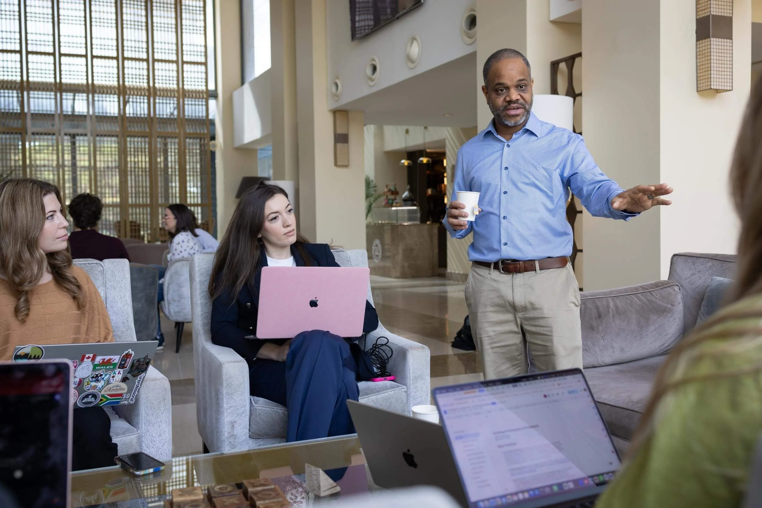 Man giving a presentation to a group of women in a modern lobby or lounge, with laptops on the table.