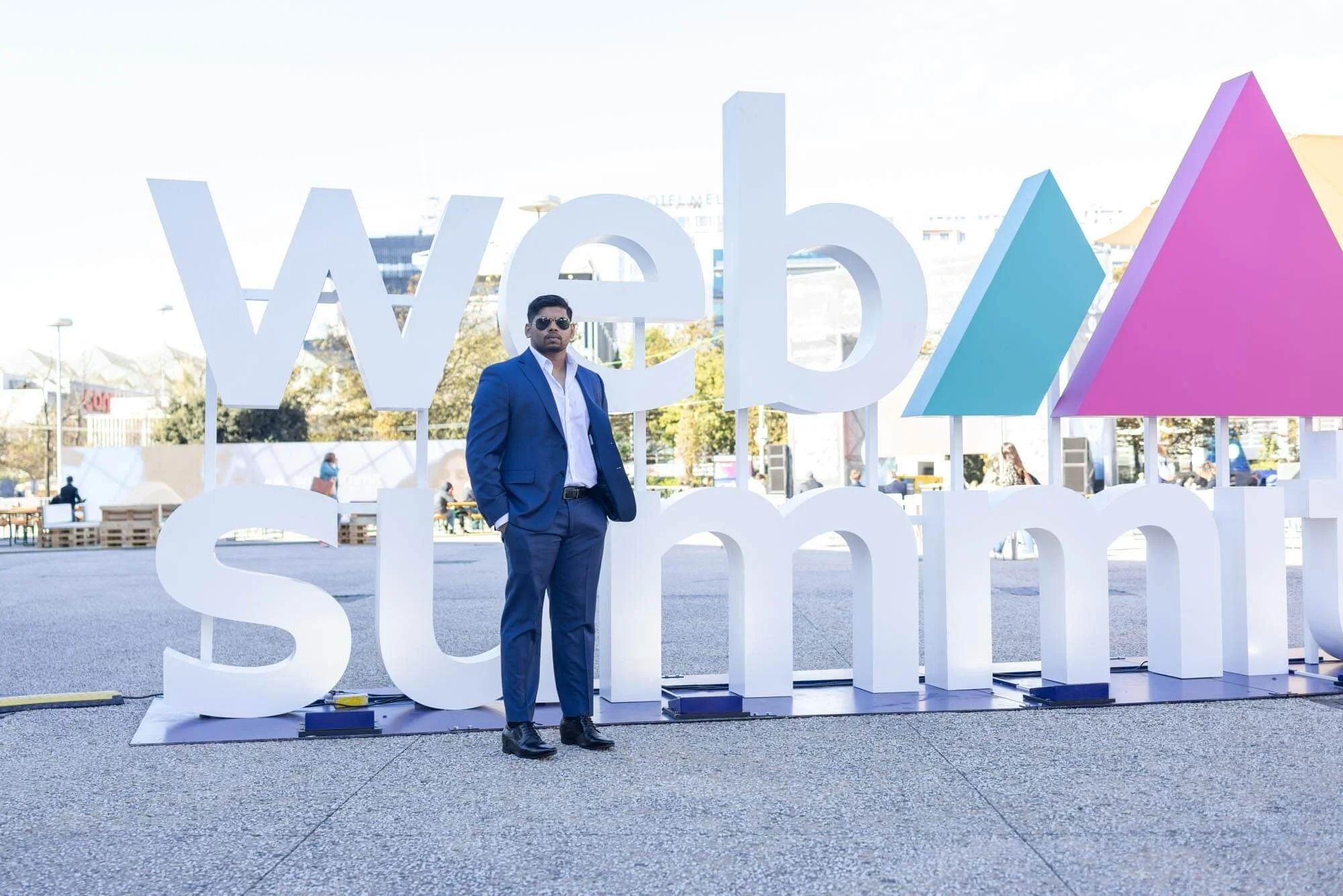 A man in a blue suit and sunglasses standing in front of large white 'web summit' sign with colorful geometric shapes in the background.