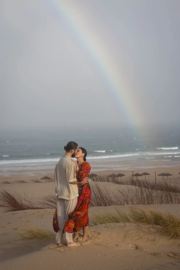 A couple kissing on a sandy beach of Cascais with a rainbow in the sky and ocean waves in the background.