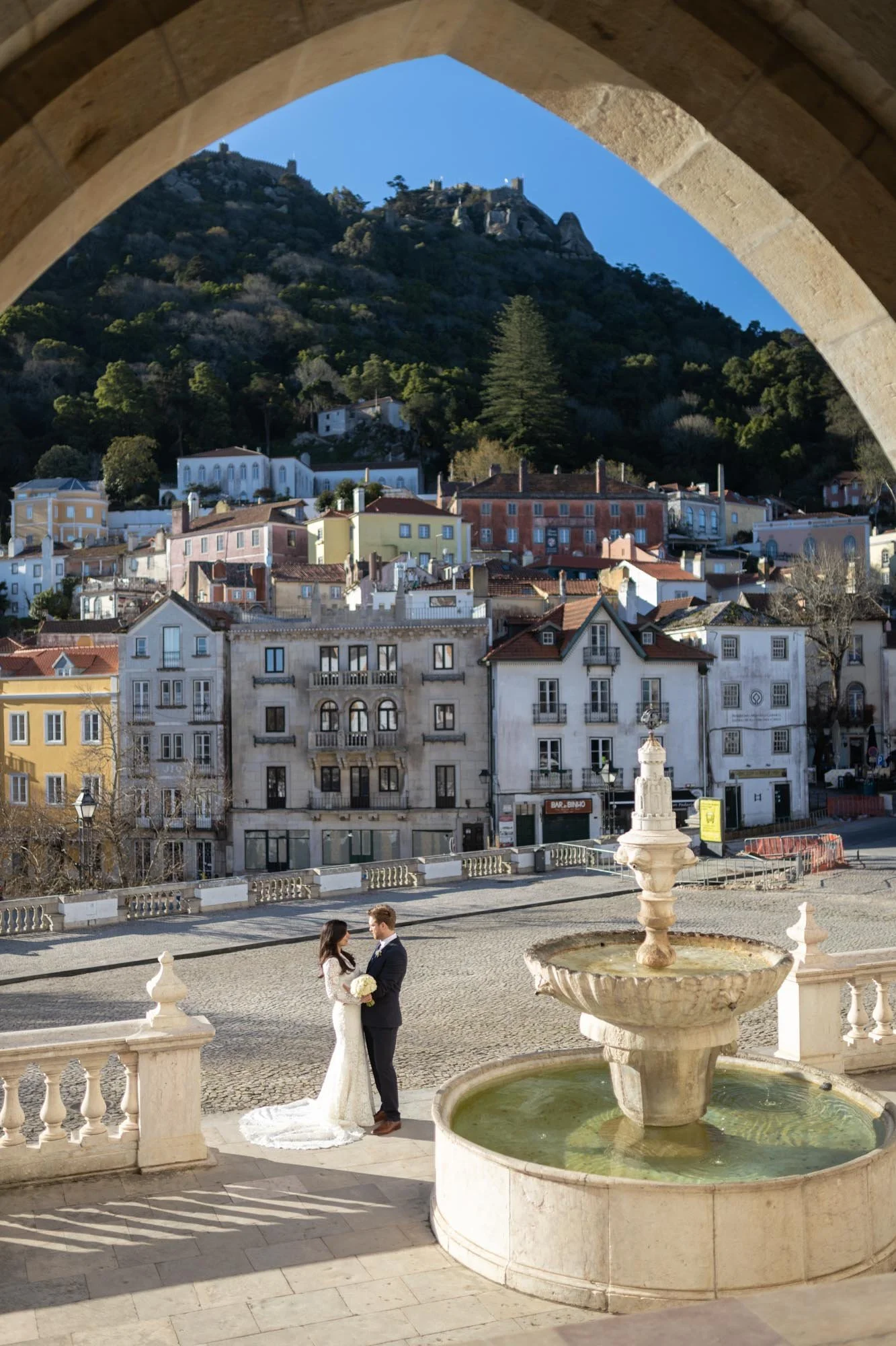 A bride and groom in wedding attire standing near a fountain in a European city square, with colorful buildings and a hillside with a fortress in the background.