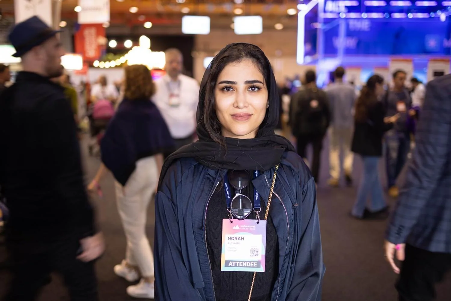 A young woman with dark hair and brown eyes smiling at a conference or event, wearing a black headscarf, black top, and a navy jacket, with an identification badge hanging from her neck that reads "Norah" and "ATTENDEE." She is in a crowded indoor ve