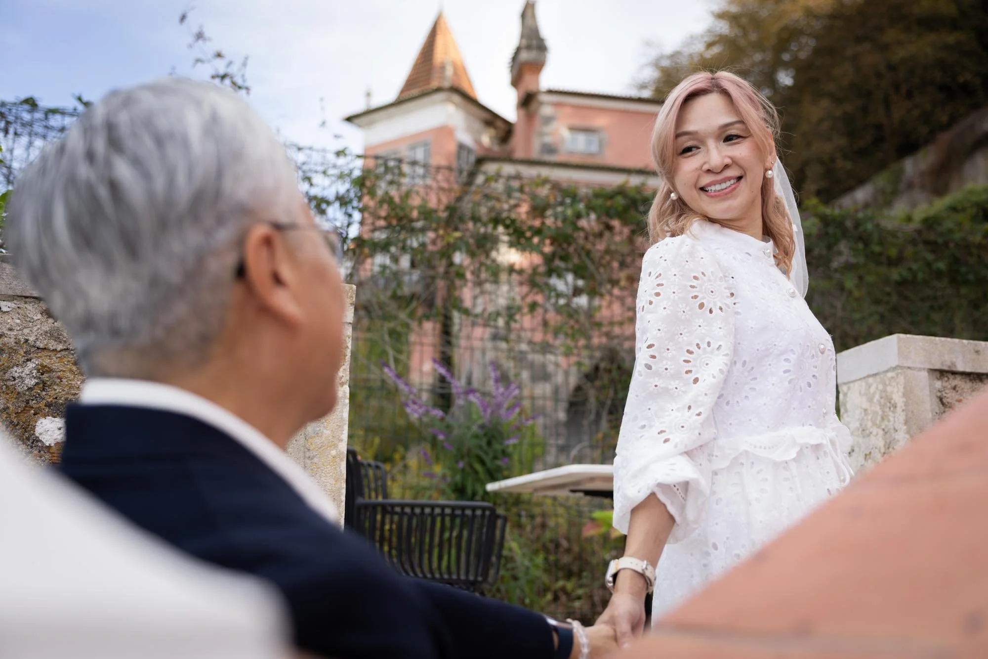 A smiling woman in a white dress with eyelet patterns, holding hands with a man with gray hair, outdoors with a house and trees in the background.