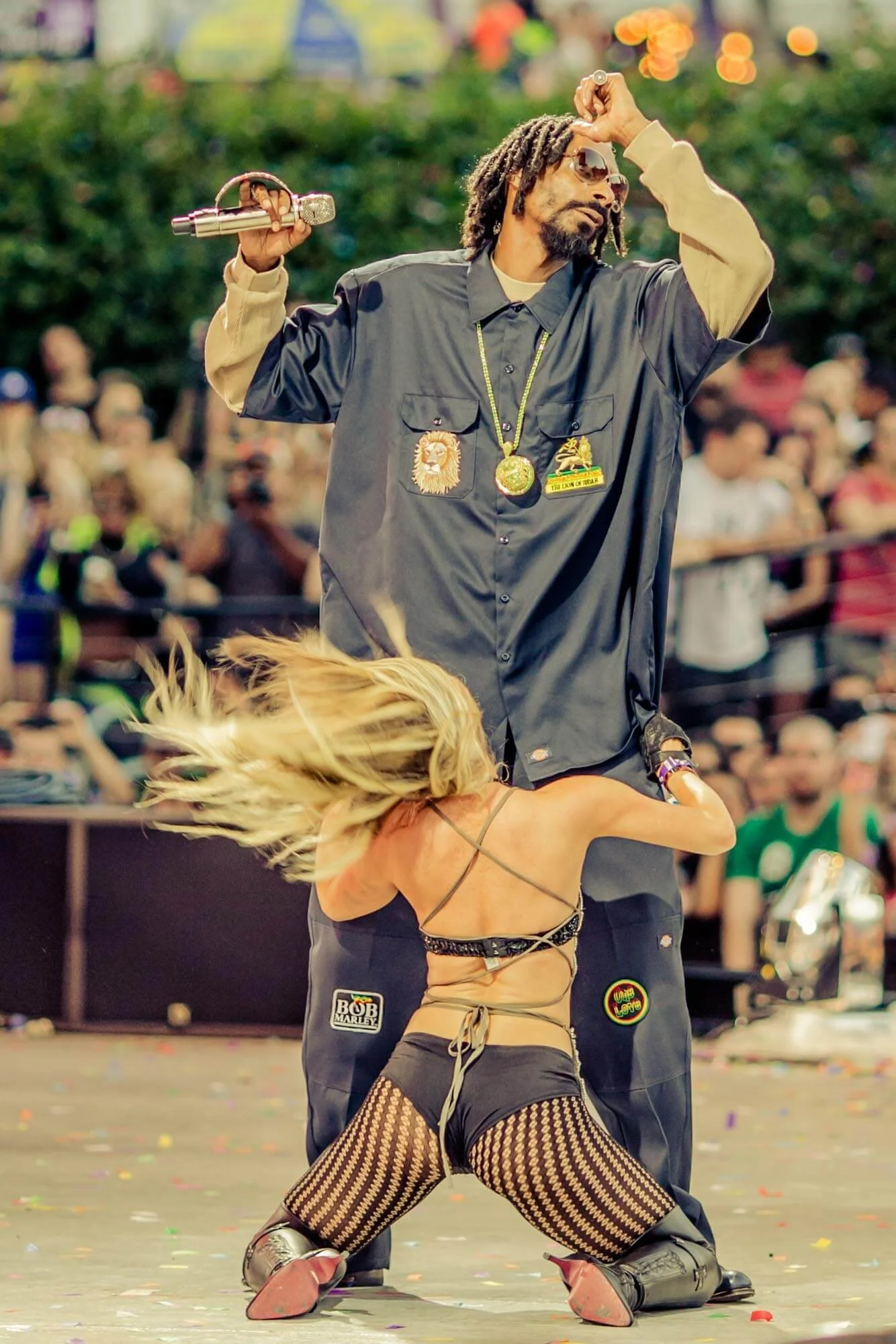 A male performer with dreadlocks, sunglasses, and a beige and black jacket holding a microphone in the air, while a female dancer with long blonde hair, fishnet stockings, and black shorts dances in front of him on stage during a lively outdoor event