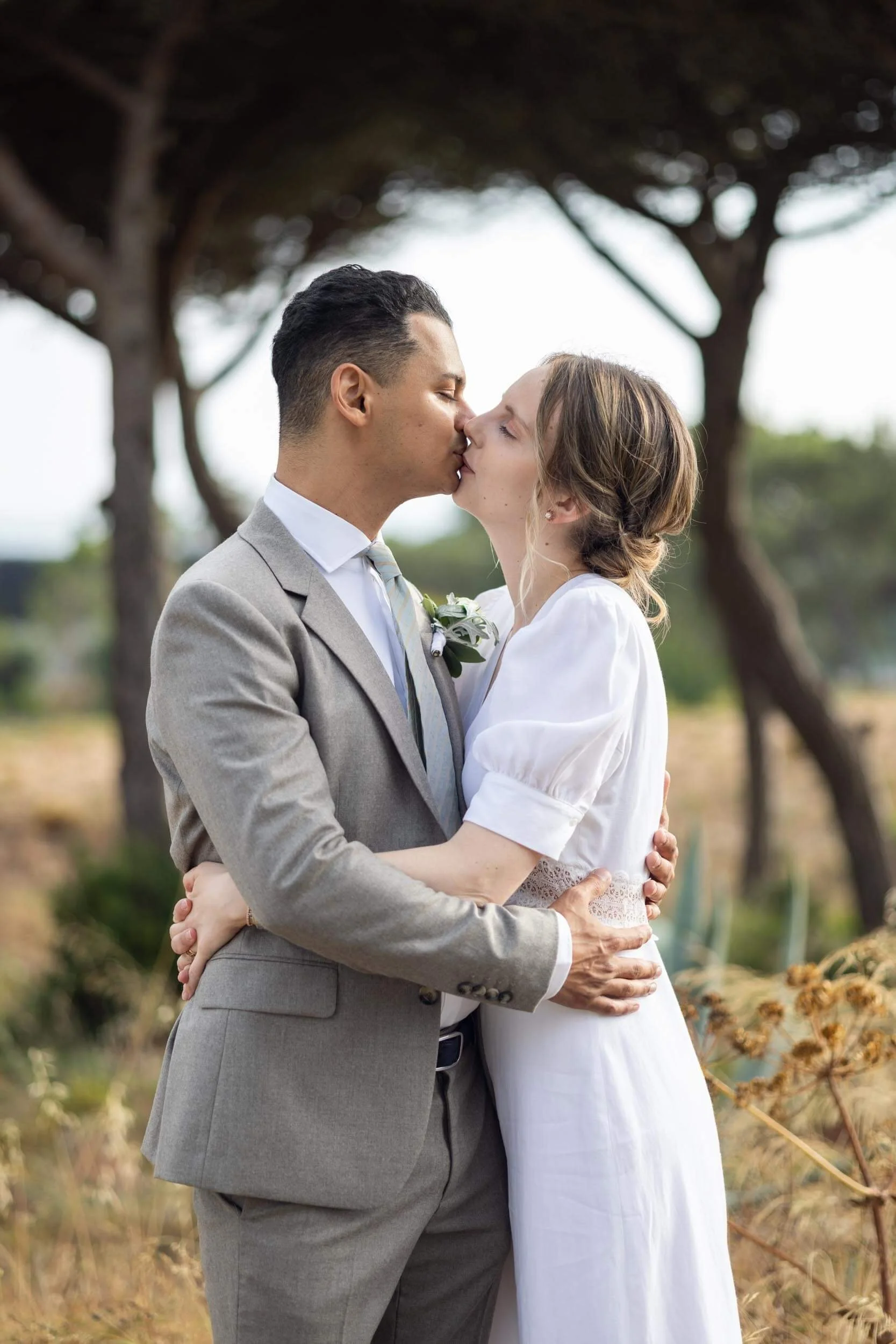 A couple dressed in wedding attire sharing a kiss outdoors with trees and dried plants in the background.
