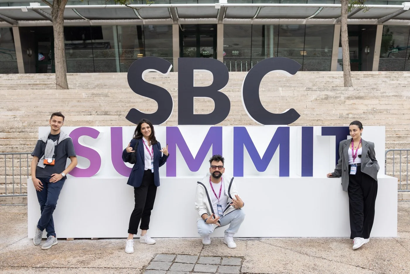 Four people standing and sitting in front of a large sign that reads 'SBC SUMMIT'. The sign has large black letters for 'SBC' and purple and white block letters for 'SUMMIT'. They are outside on steps near a building with glass windows and trees.