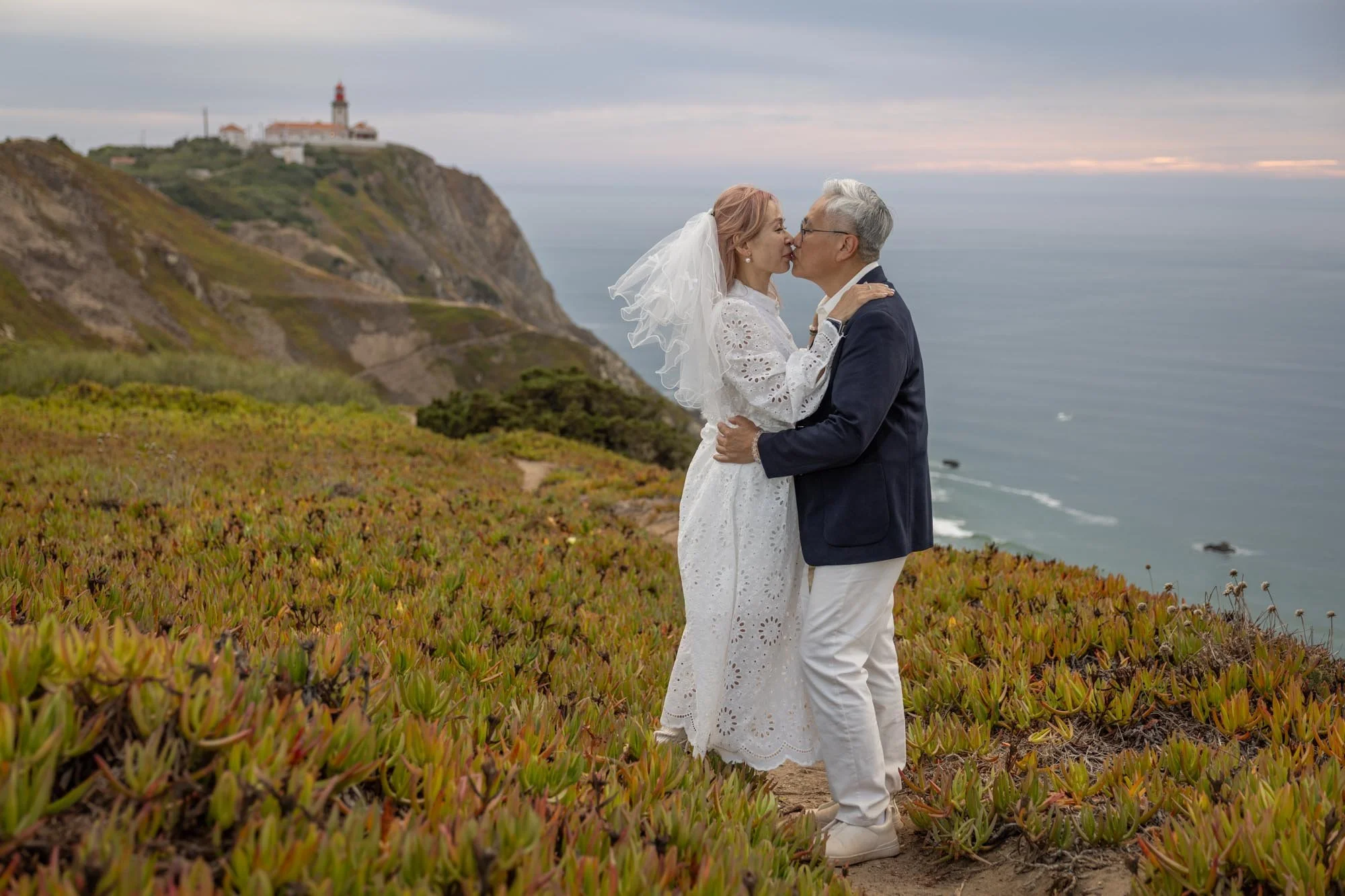 A couple in wedding attire sharing a kiss on a cliffside overlooking the ocean, with a lighthouse on a hill in the background.