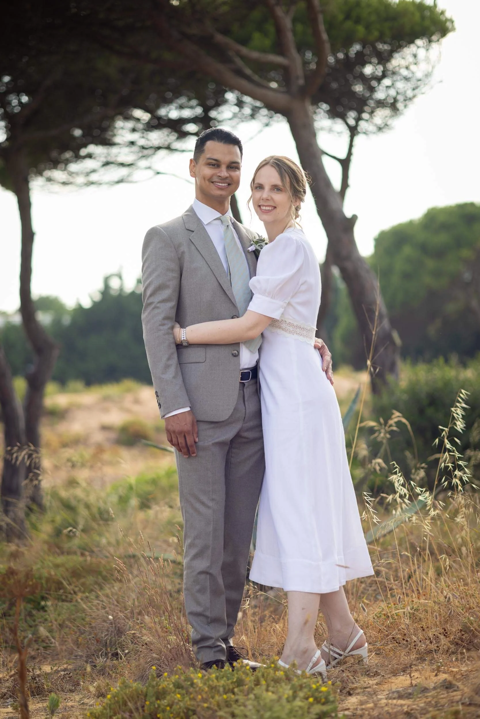 A couple dressed in wedding attire, standing outdoors on a grassy area with trees in the background, smiling and embracing each other.