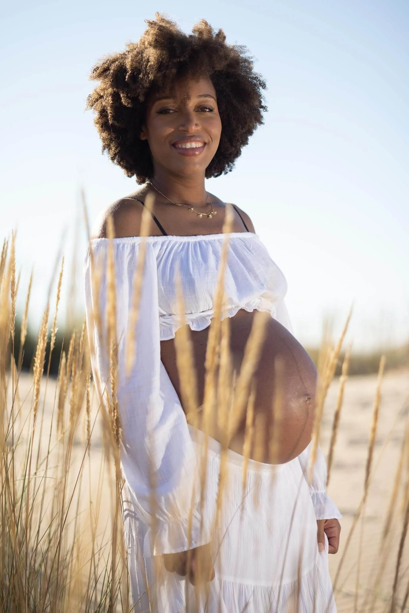 A pregnant woman with curly hair smiling outdoors in a field of tall grass, wearing a white off-the-shoulder top and white skirt.