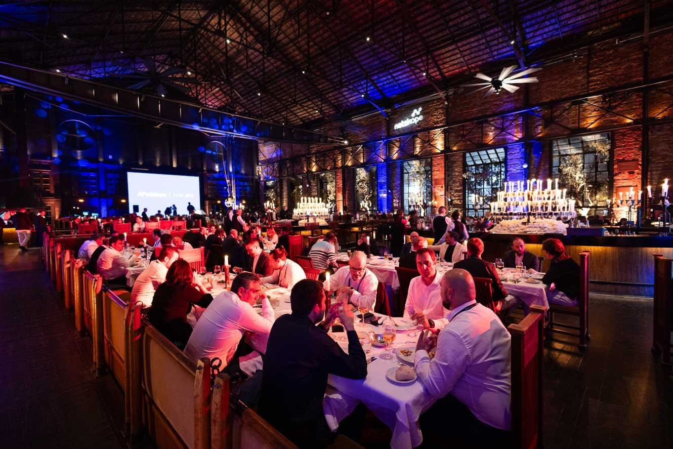 People dining at long tables in a large, industrial-style event space with brick walls, high ceilings, and colorful lighting, celebrating with a large cupcake display and candles.